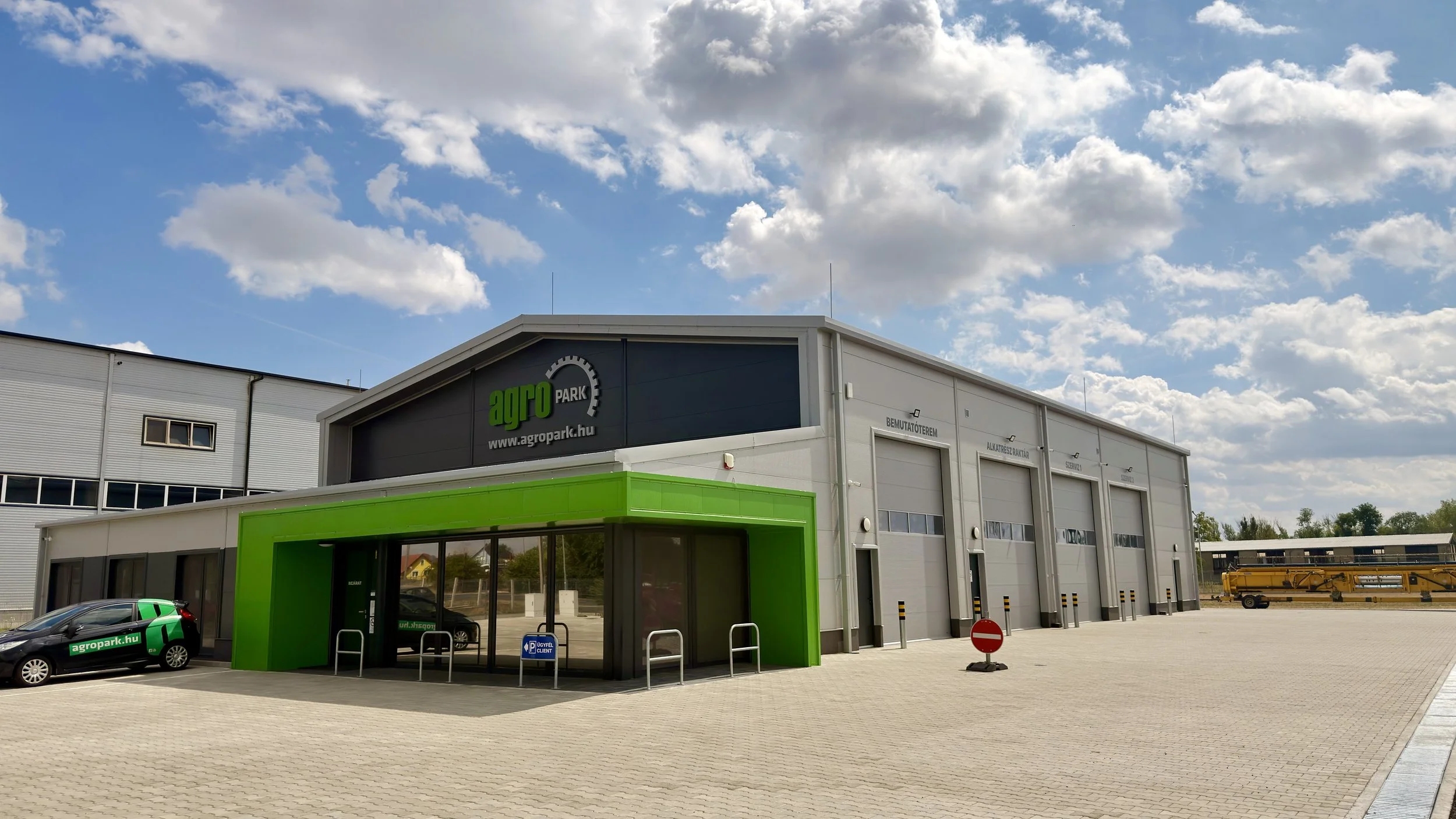 Exterior of an agricultural equipment store named Agro Park with a modern gray and green facade, large garage doors, a parking lot with a black vehicle marked 'agropark.hu', under a partly cloudy sky.