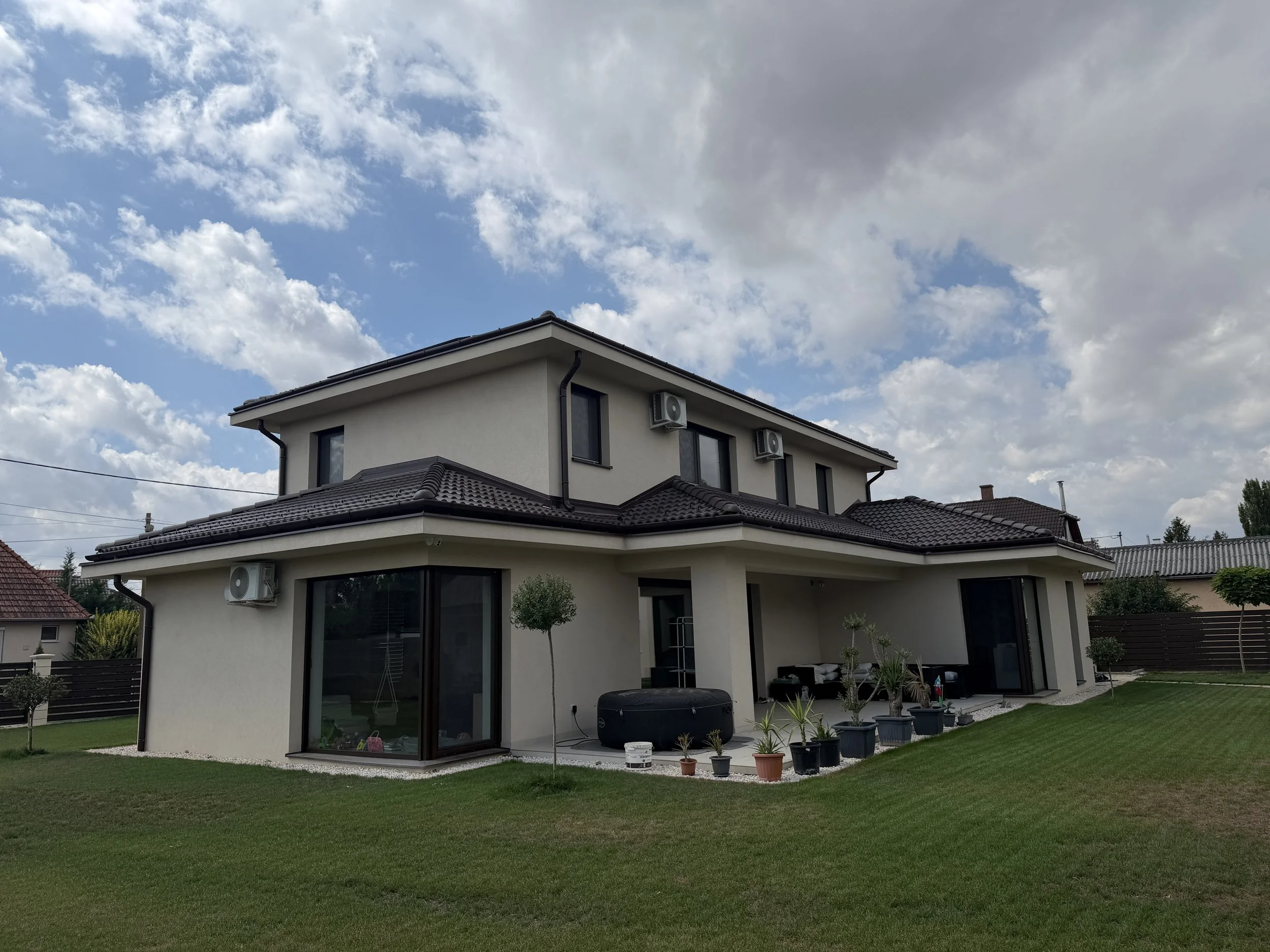 A modern two-story house with a tiled roof, multiple windows, and air conditioning units mounted on the exterior walls. The backyard has a well-maintained lawn, numerous potted plants, and some outdoor furniture, with a partly cloudy sky overhead.