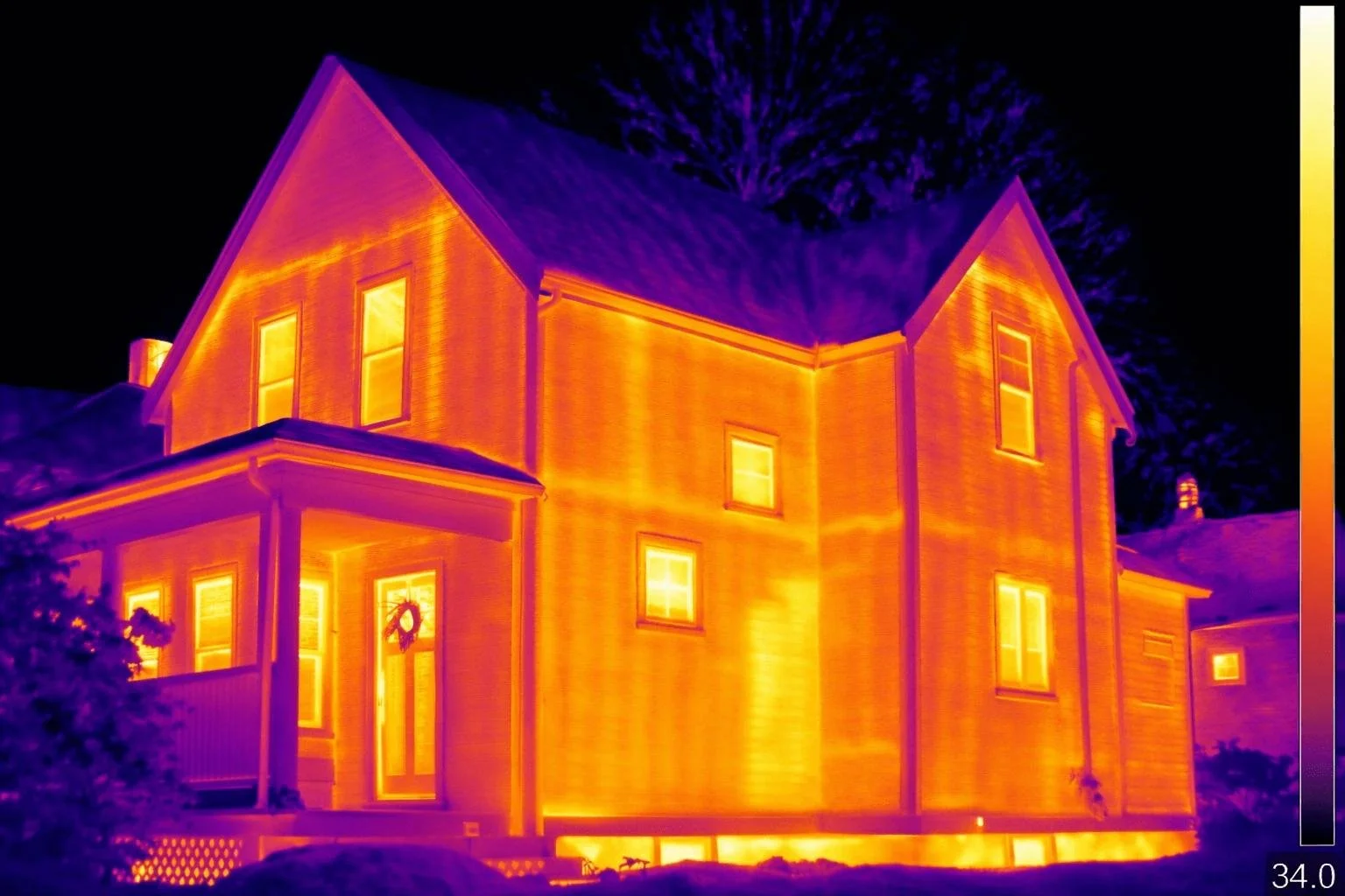 Infrared image of a two-story house with several windows, illuminated warmly from inside, with a dark night sky and a tree in the foreground.