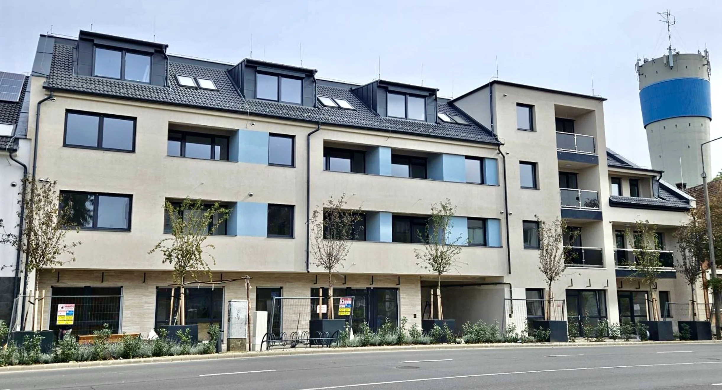 Multi-story modern residential building with beige and blue exterior, large windows, balconies, and a rooftop deck, located on a city street with trees and a water tower in the background.