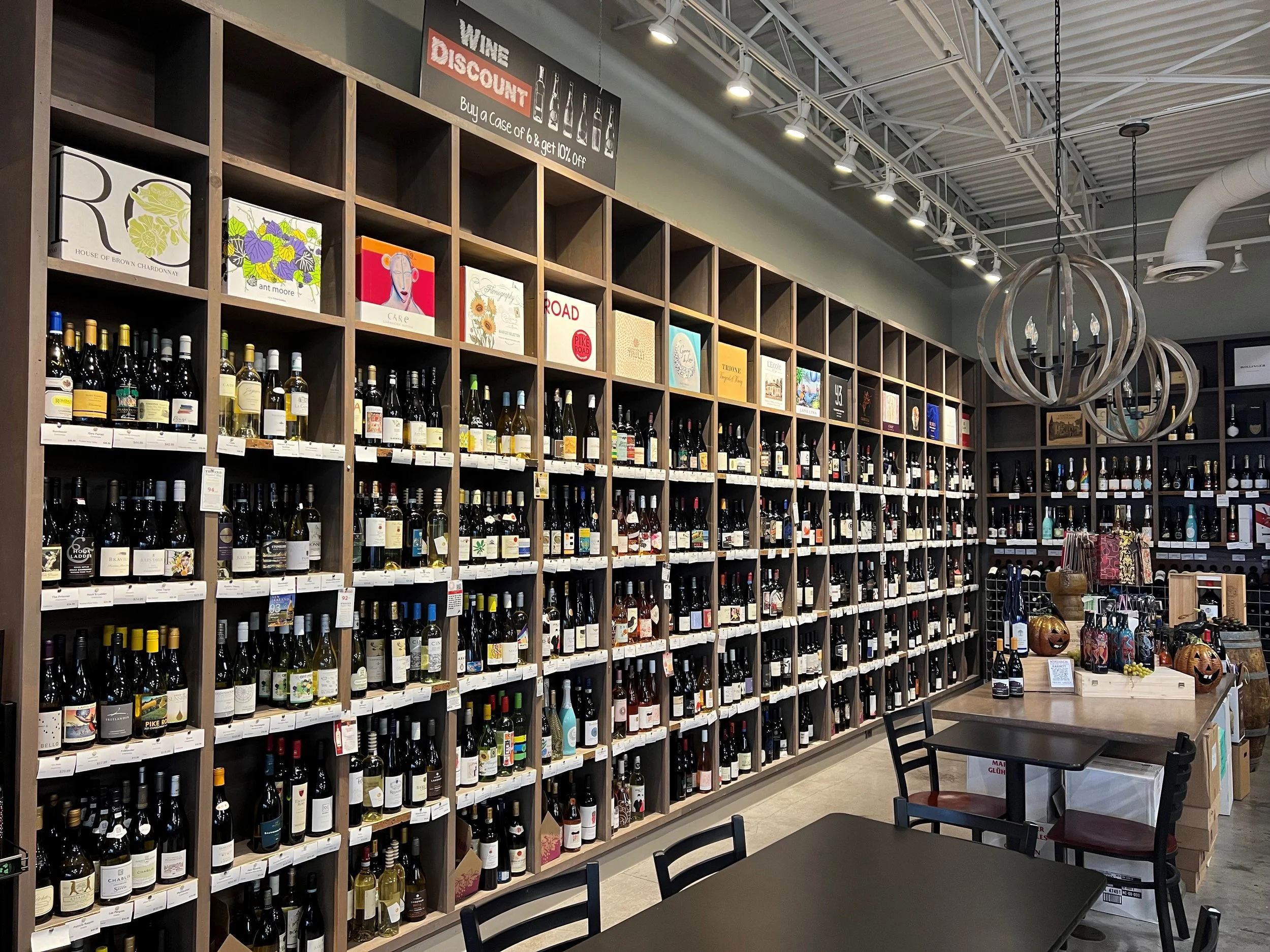 Interior of a retail store displaying shelves stocked with bottles of wine and assorted products, including a small table with decorative items.