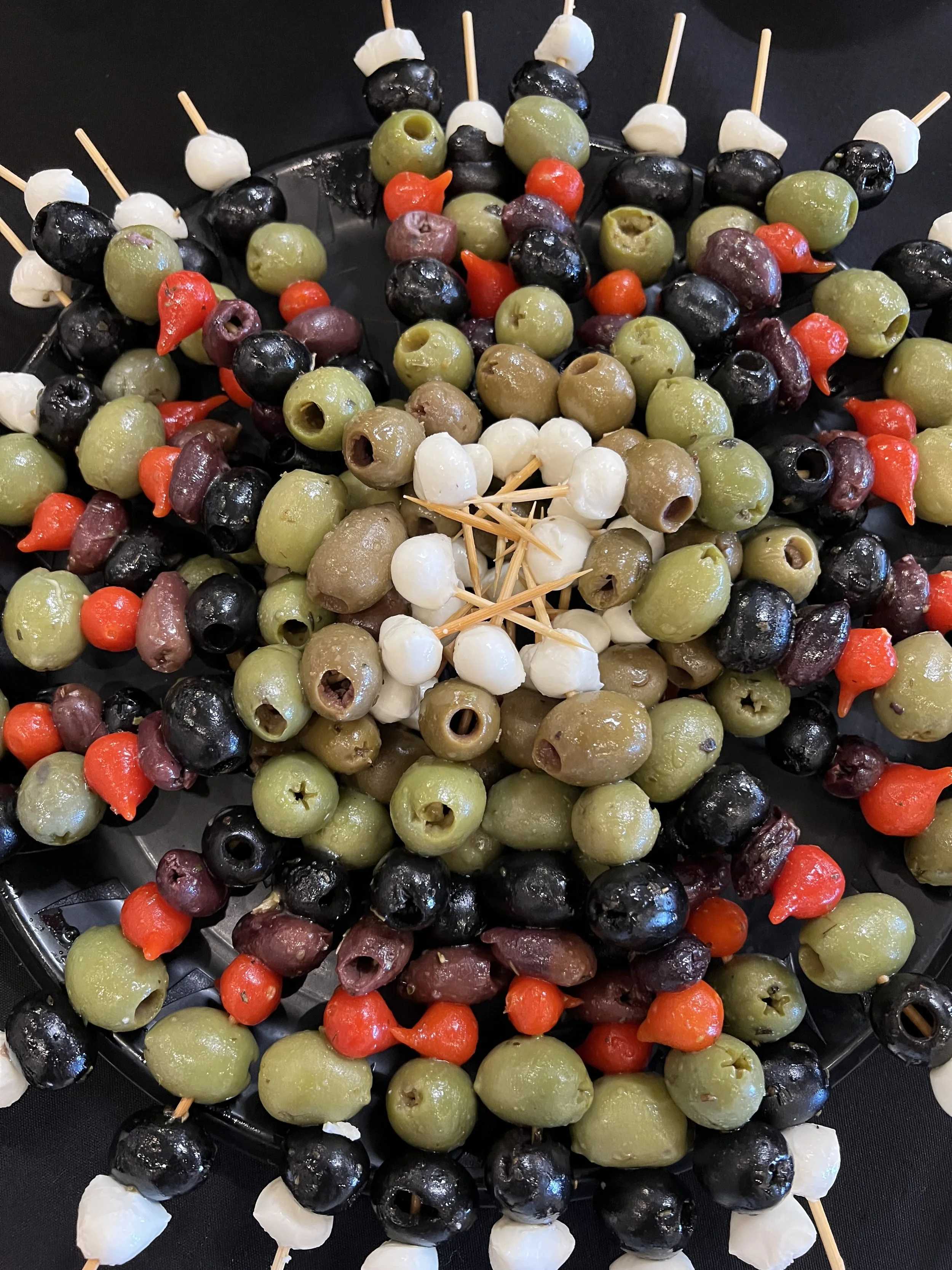 Skewers with green, black, white, and red olives on a black surface.