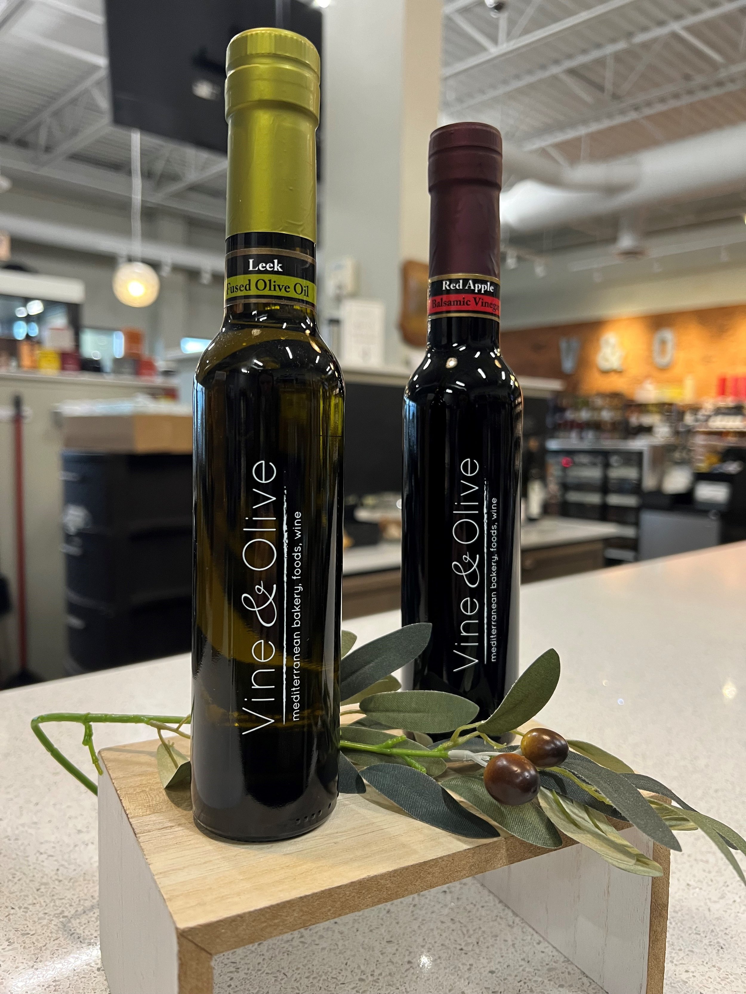 Two bottles of olive oil and balsamic vinegar placed on a wooden board with decorative green leaves and brown olives, set on a white table in a restaurant or store.
