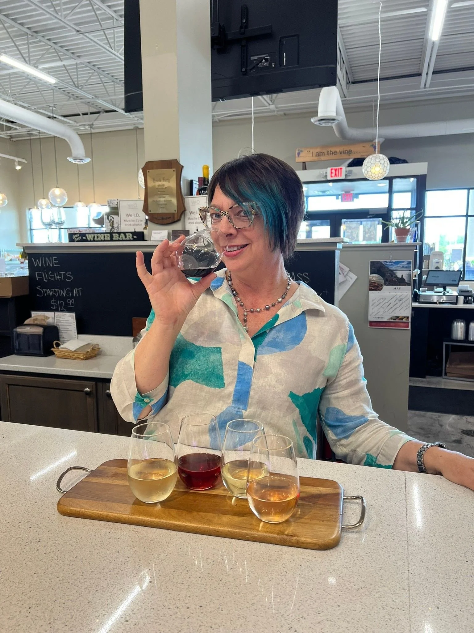 Woman with short dark hair and colorful highlights, wearing glasses and a patterned blouse, smiling while tasting red wine in a restaurant or wine tasting room, with a flight of five glasses of wine on a wooden tray in front of her.