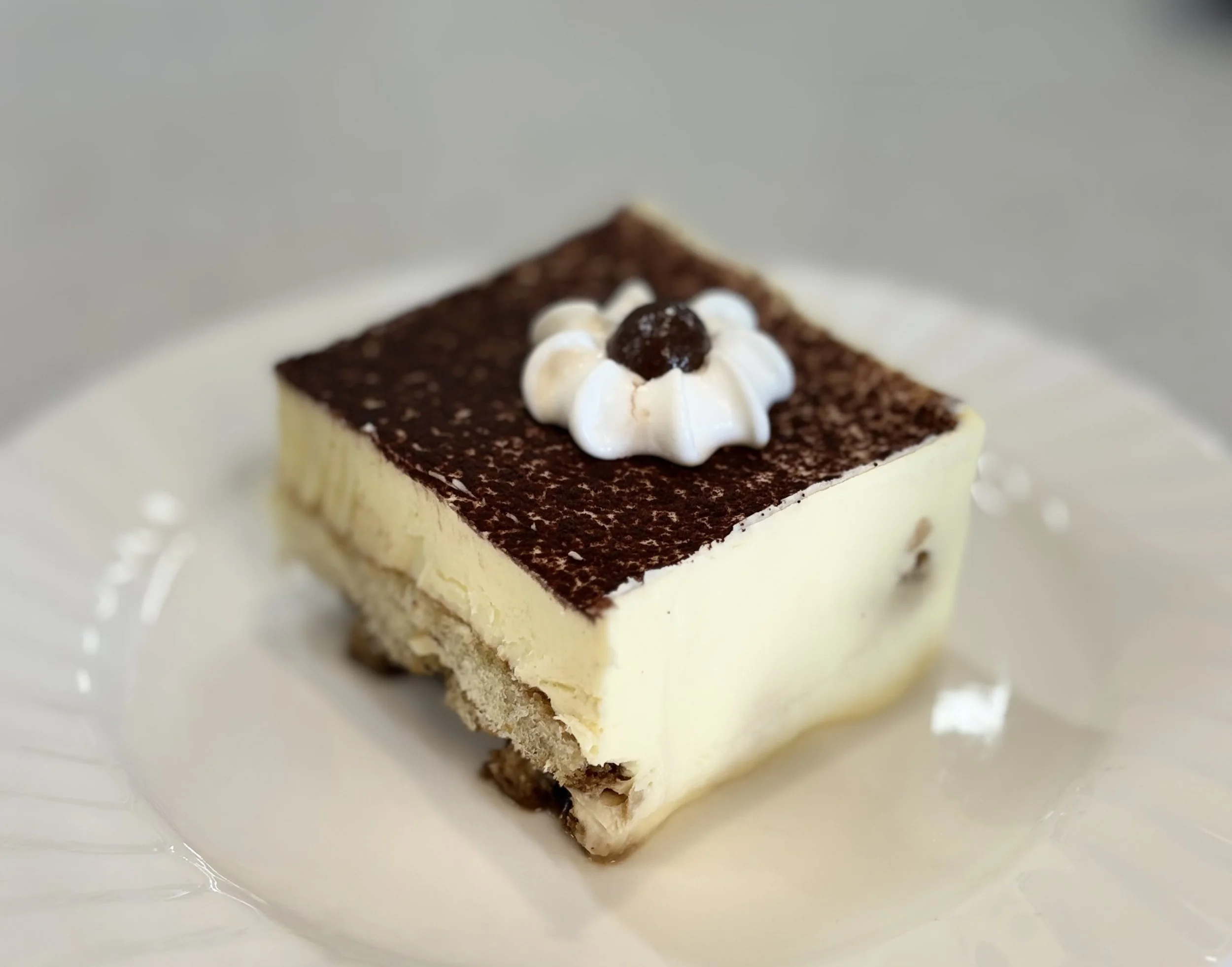 A slice of cheesecake topped with whipped cream, a cherry, and a dusting of cocoa powder, served on a white plate.
