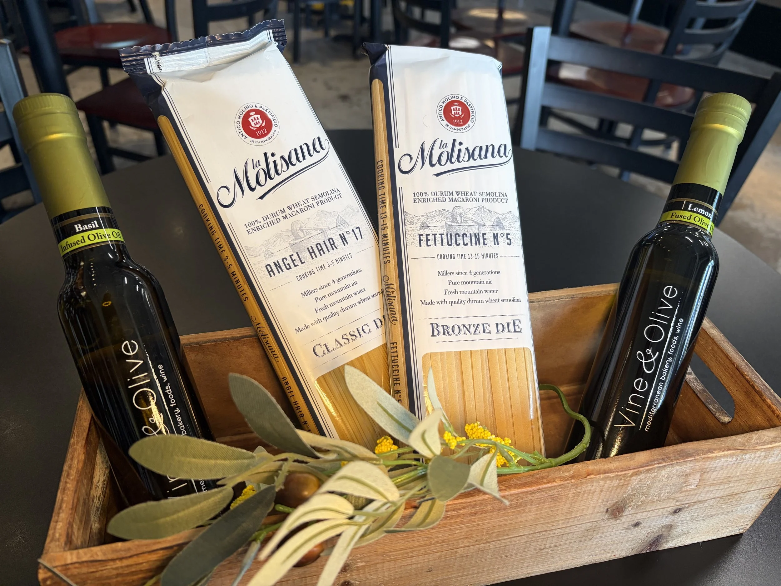 Two packets of pasta, one labeled Angel Hair number 17 and the other Fettuccine number 5, are placed in a wooden basket along with two bottles of infused olive oil, one labeled basil and the other lemon.