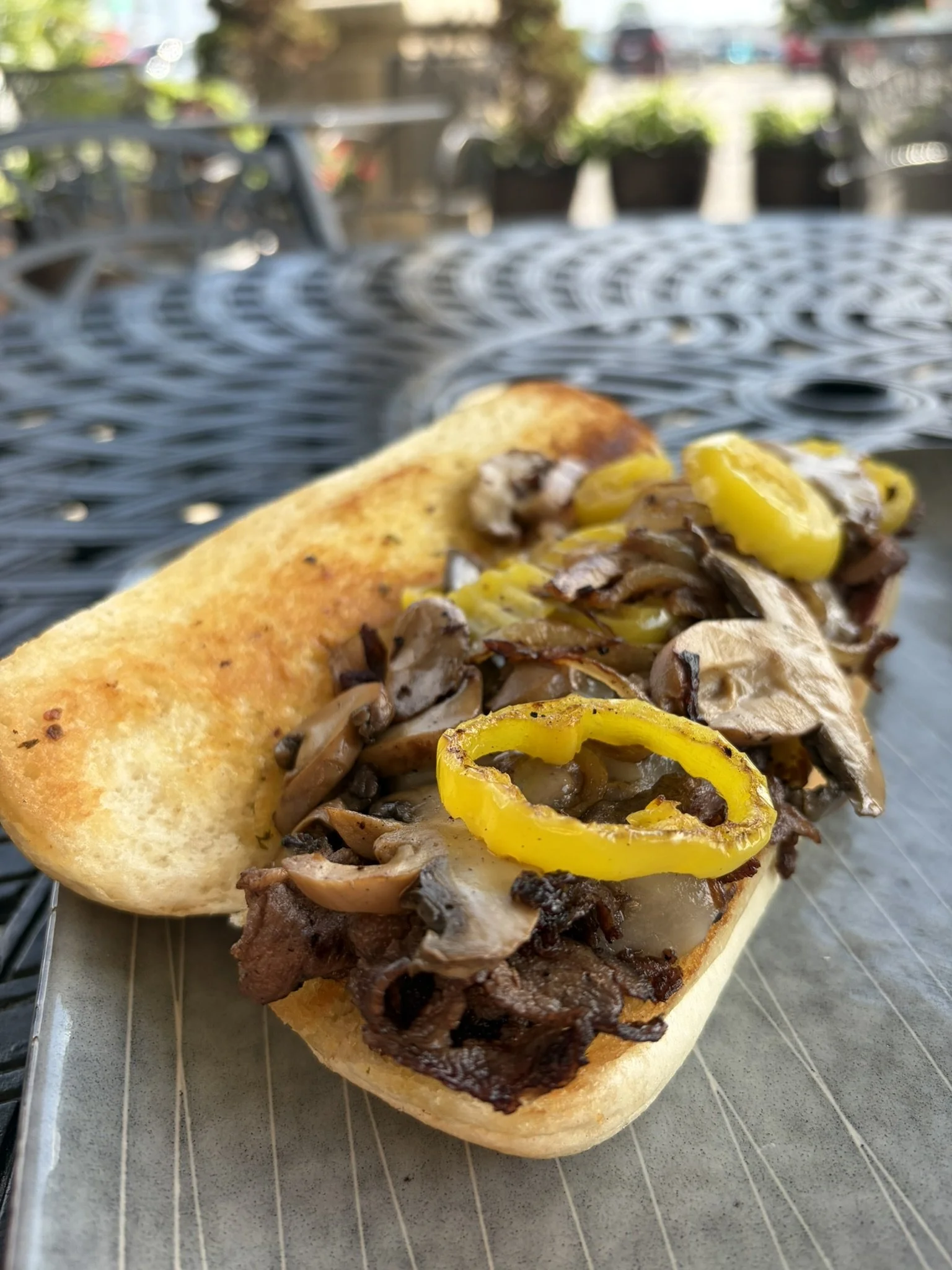 Philly cheesesteak sandwich with sliced yellow peppers and mushrooms on a hoagie bun, resting on a metal tray.
