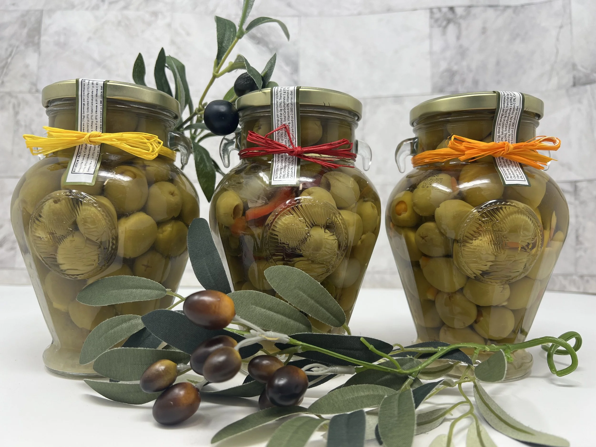 Three glass jars with gold lids filled with green olives, decorated with colorful ribbons, and a sprig of olives and leaves in front. A branch with black and purple olives and green leaves is also present.