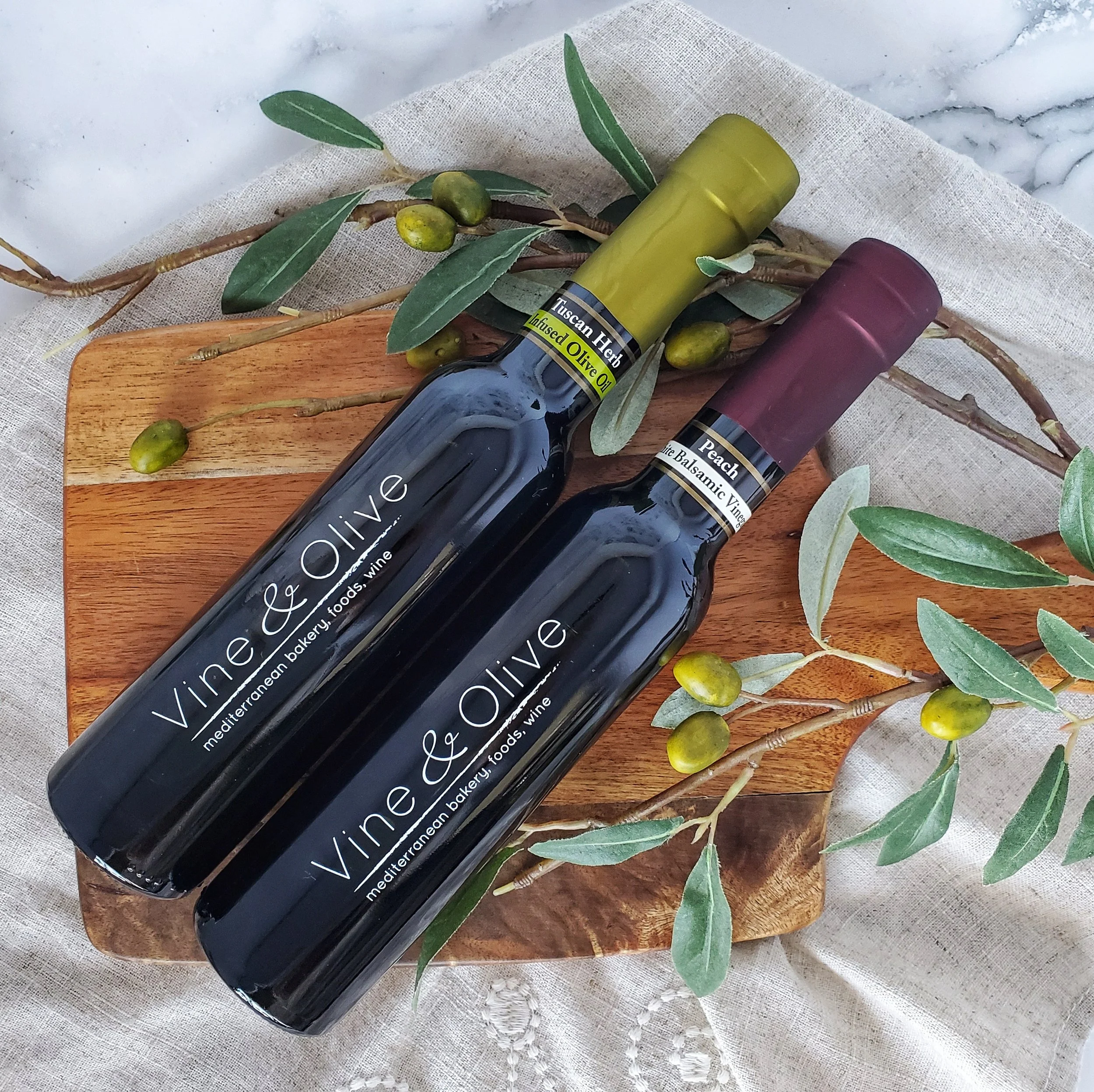Bottles of organic olive and peach balsamic vinegar on a wooden board surrounded by olive branches and leaves, with a white fabric background.