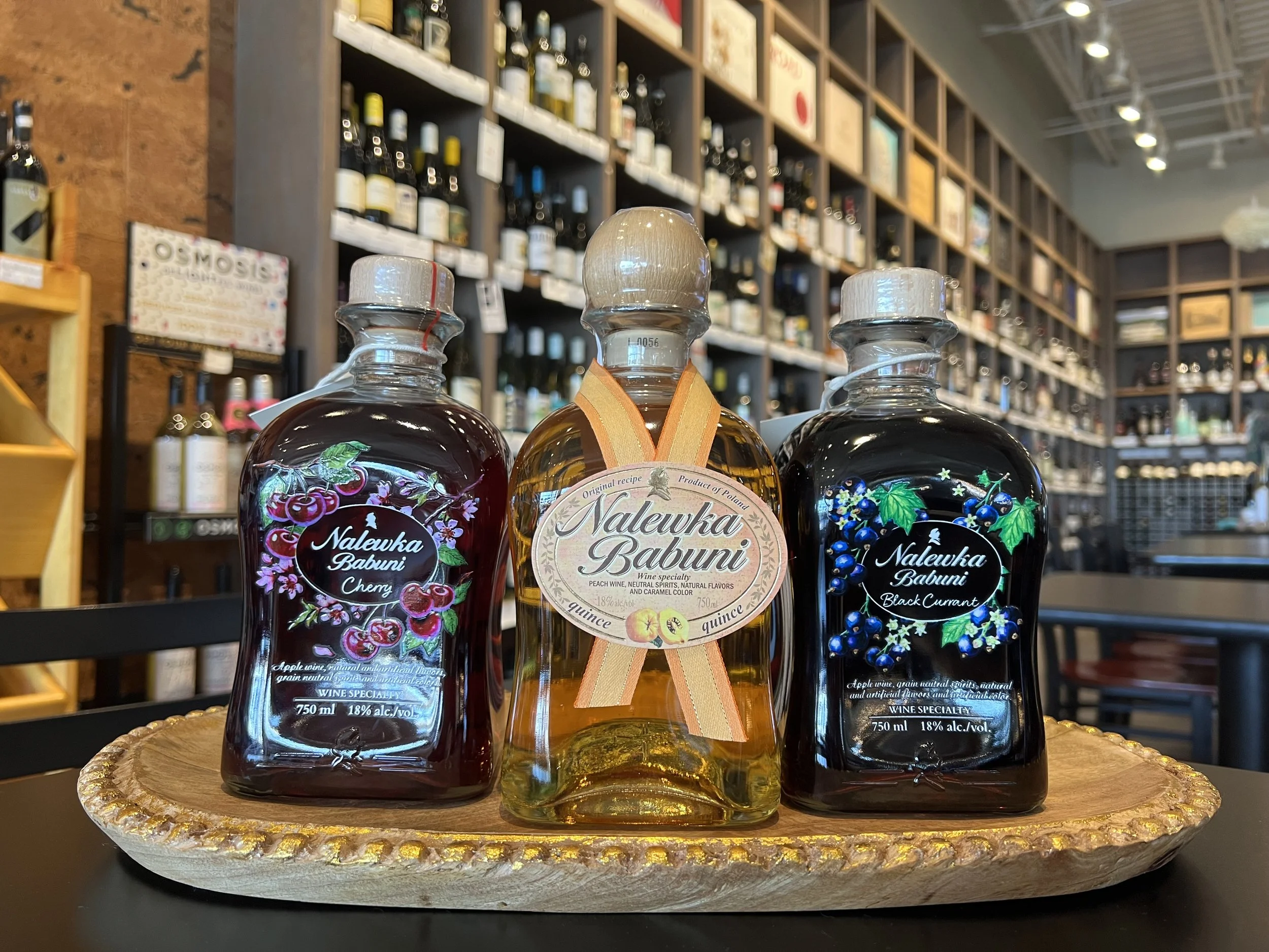 Three bottles of Nalewka Babuni liquor on a decorative tray, with a blurred background of store shelves filled with bottles and boxes.