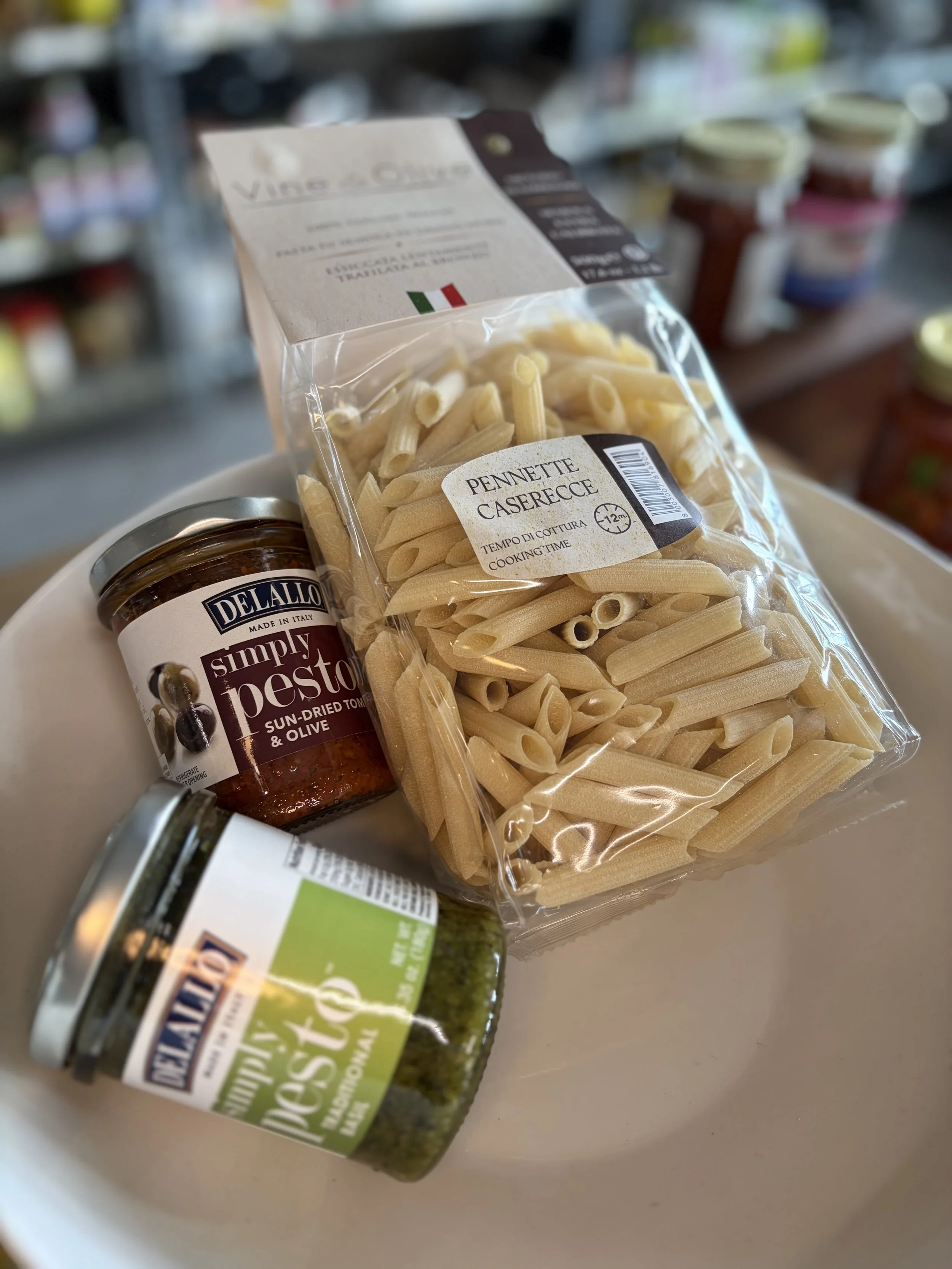 A package of penne caserecce pasta with Italian flag on the label, a jar of Simply Pesto sun-dried tomato and olive spread, and other jars of food on a store shelf in the background.