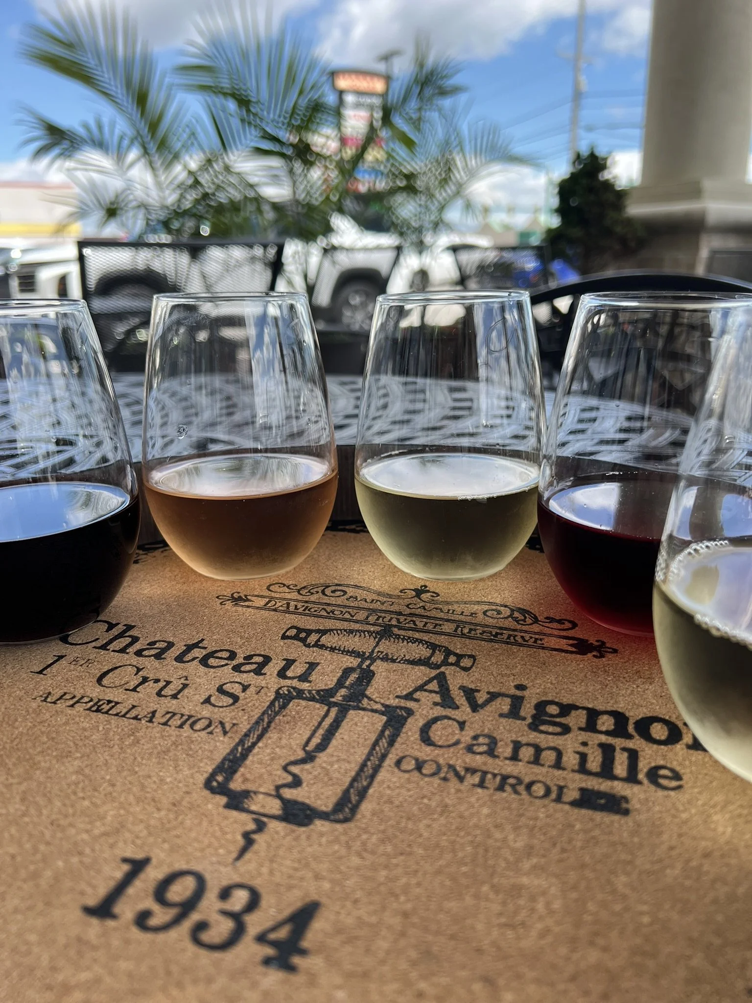 Four wine glasses filled with different colored wines on a table at an outdoor restaurant, with palm trees, a signboard, and a blue sky in the background.