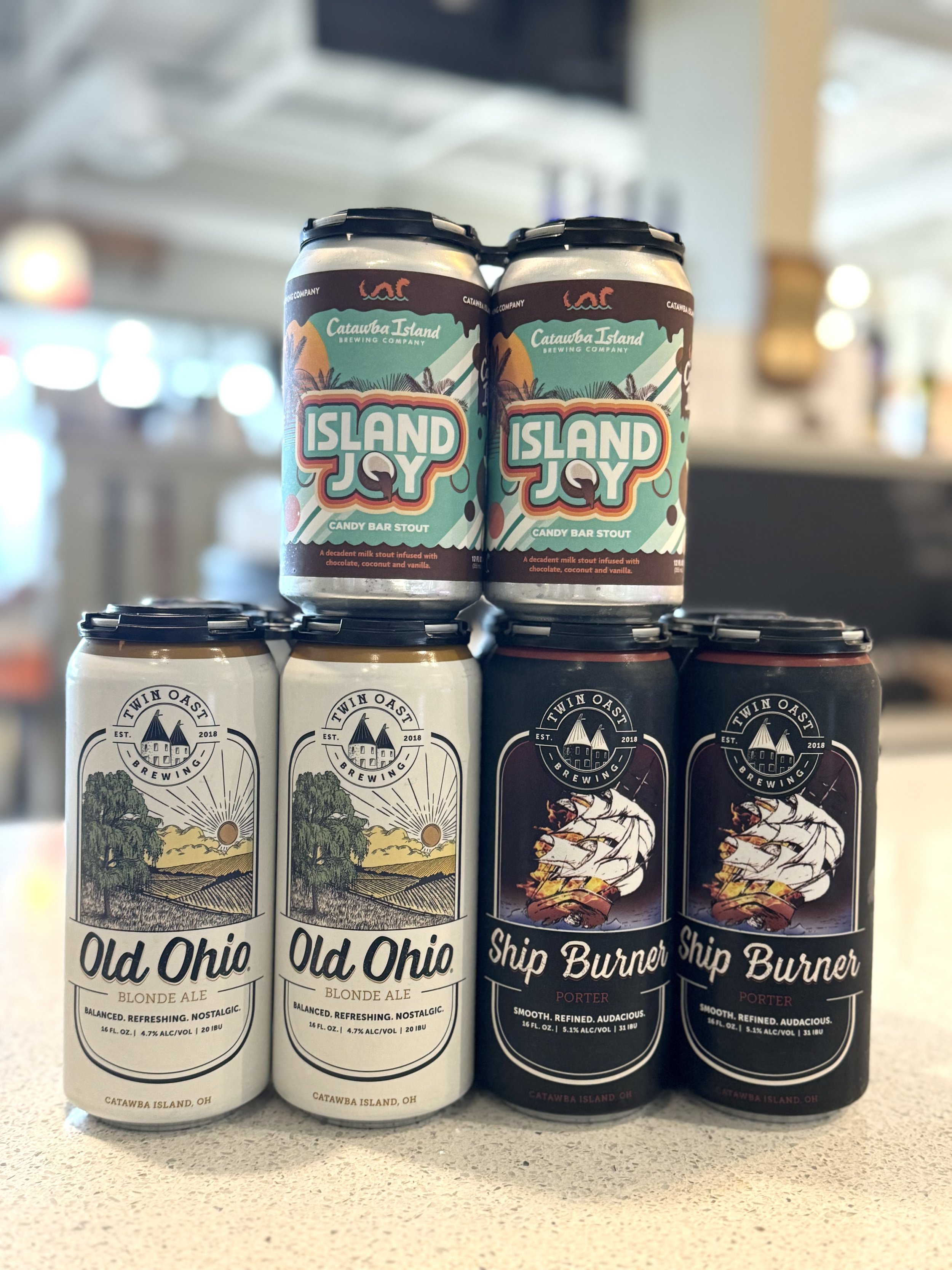 Three cans of beer stacked on a table, with a blurred background. The cans have colorful labels, with two labeled 'Old Ohio Blonde Ale' and one labeled 'Ship Burner Porter' from Catawba Island Brewing Company.