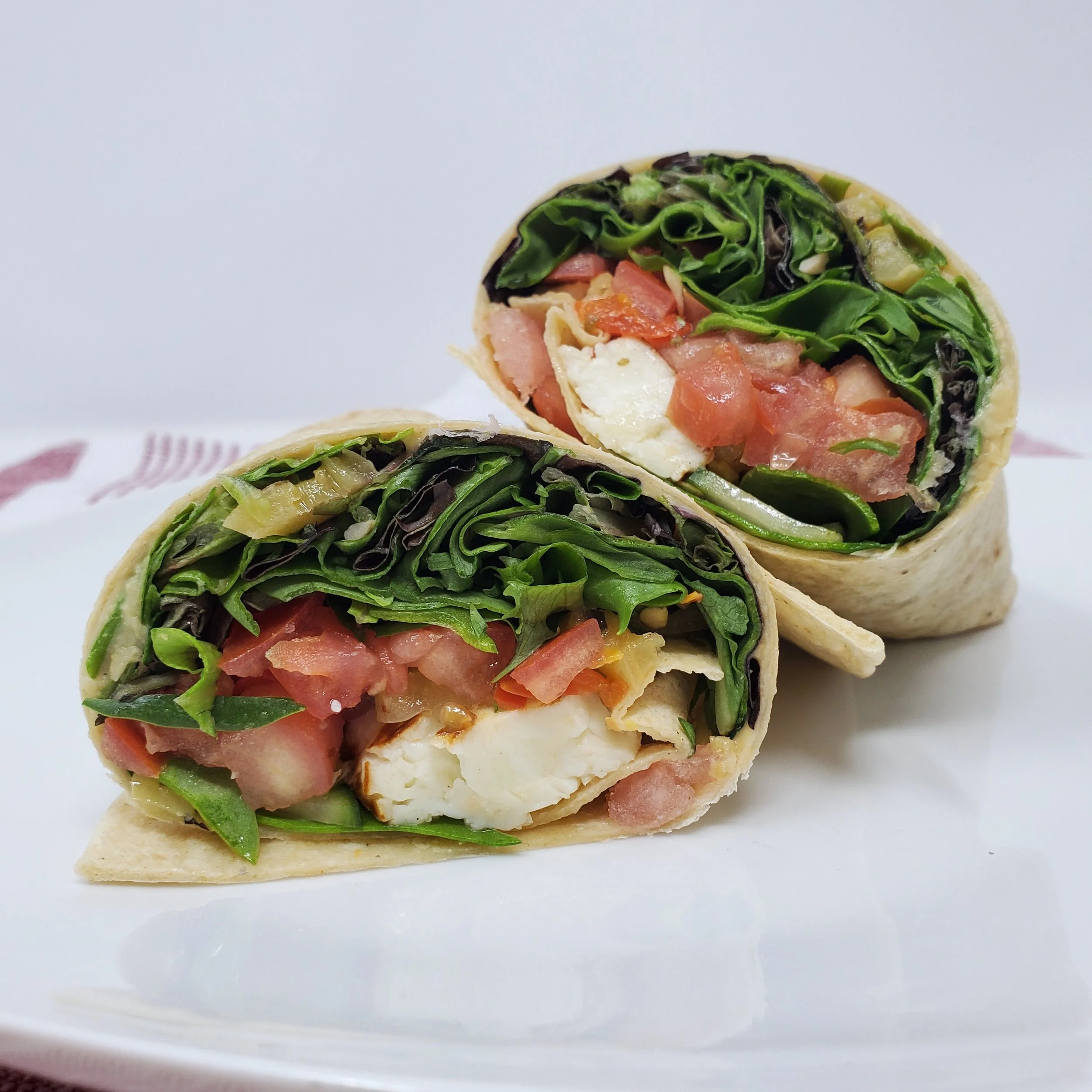 Close-up of a wrap sandwich filled with spinach, tomatoes, cheese, and ham on a white plate against a white background.