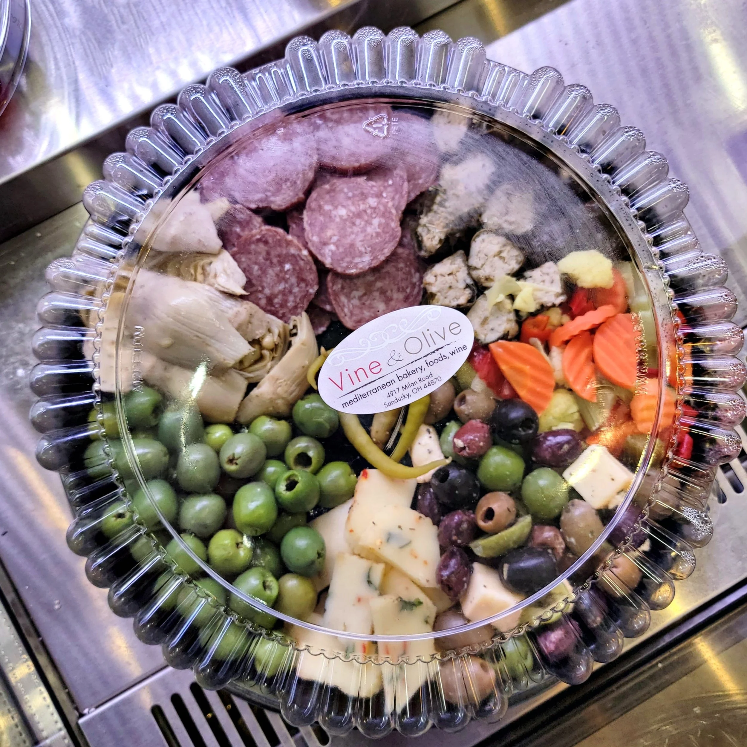 A clear plastic platter with various types of cheese, green olives, and slices of salami on a stainless steel surface.