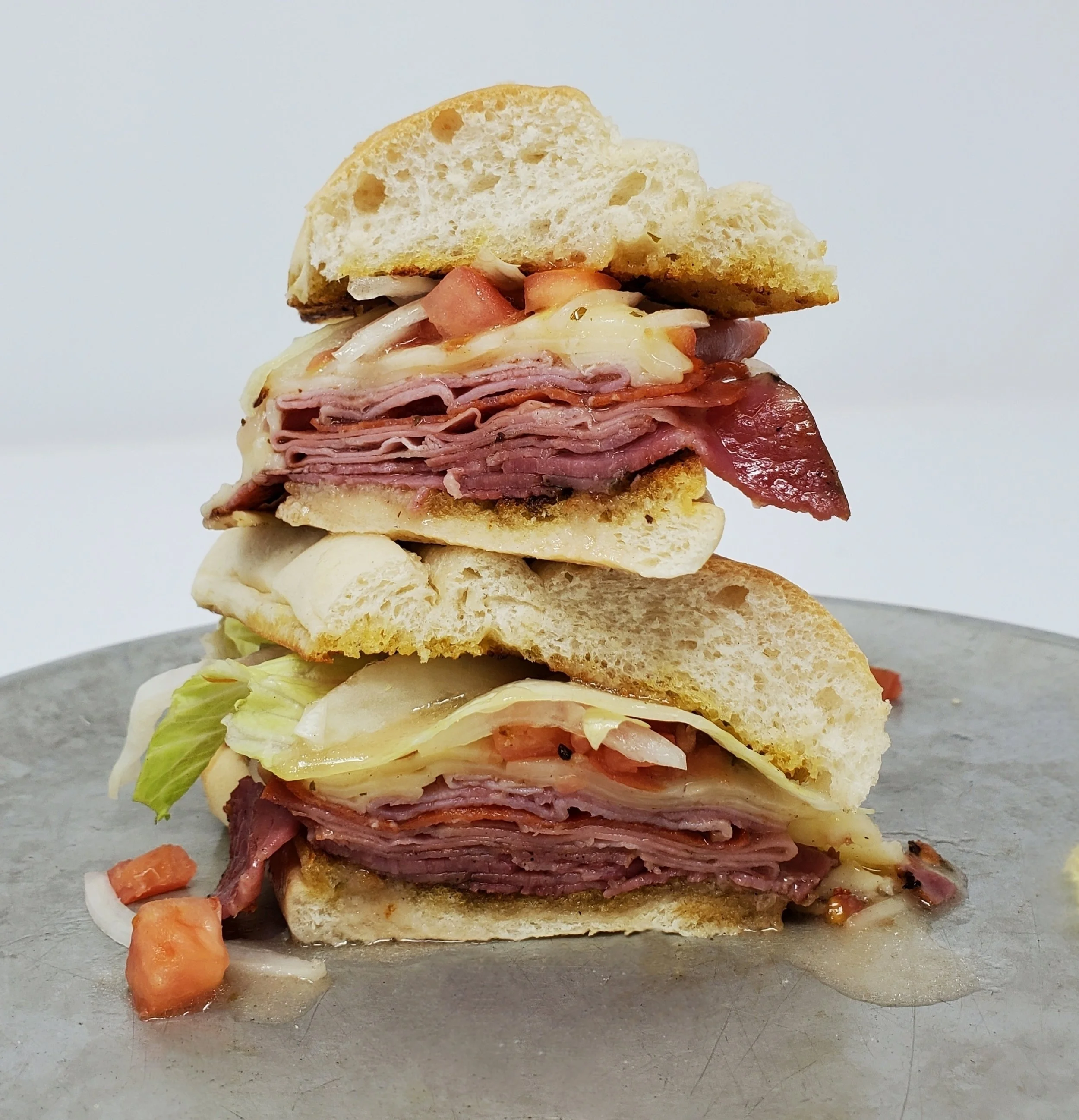 Close-up of a multi-layered sandwich with deli meats, cheese, lettuce, tomato, and bread, placed on a gray surface against a plain white background.
