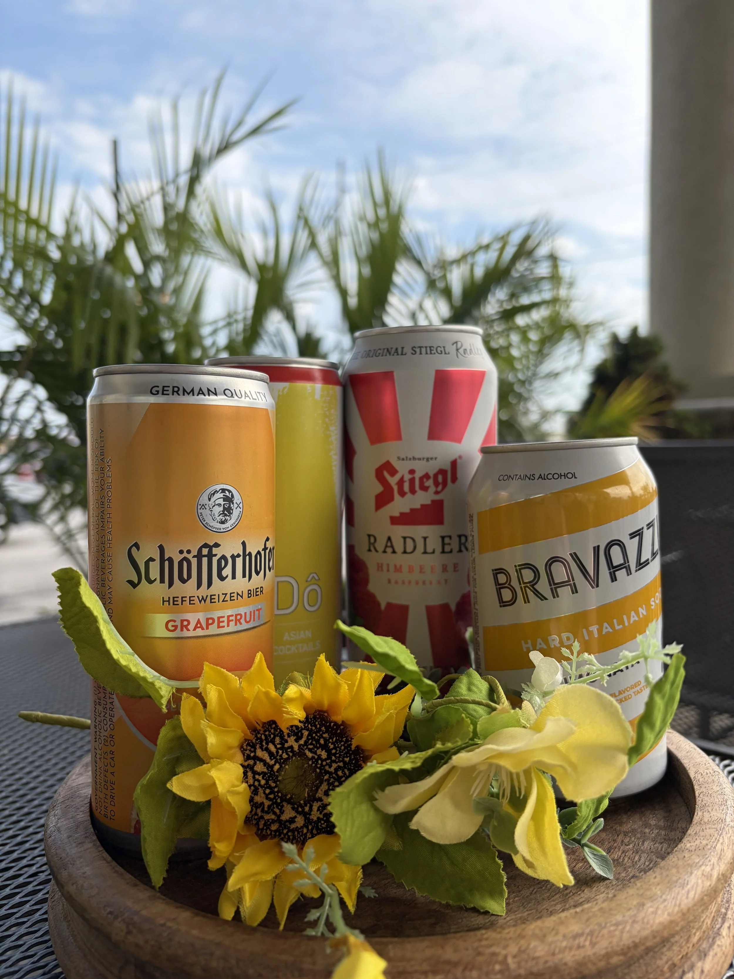 A wooden bowl with assorted colorful beverage cans and yellow flowers on a dark outdoor surface with a cloudy sky and palm trees in the background.
