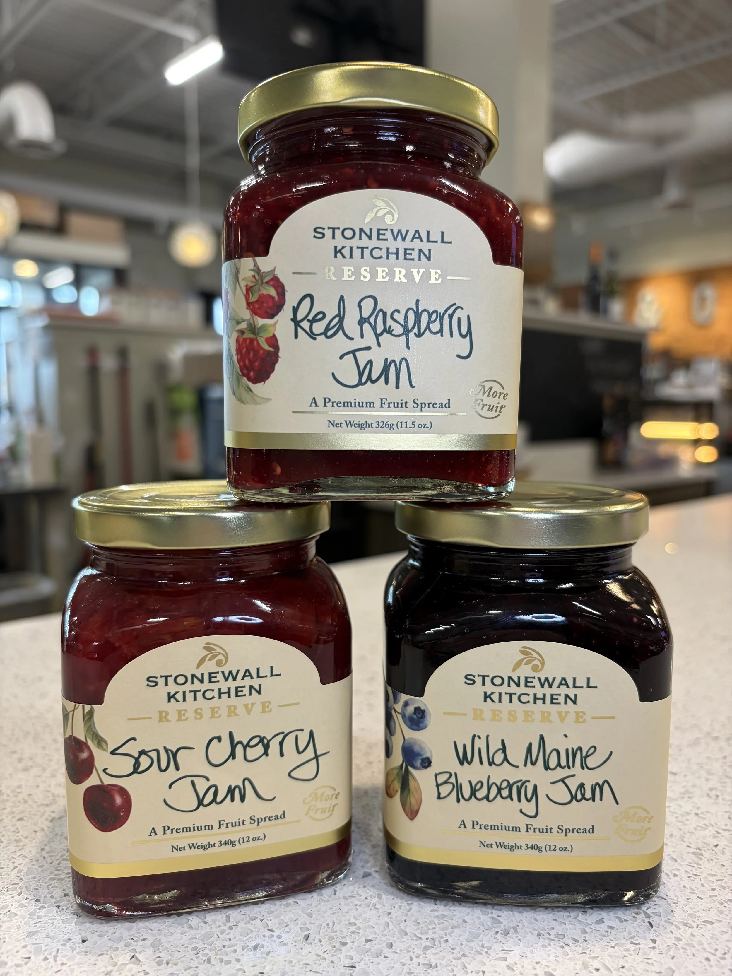 Three jars of Stonewall Kitchen preserve on a table, with the top jar labeled 'Red Raspberry Jam', the left jar labeled 'Sour Cherry Jam', and the right jar labeled 'Wild Maine Blueberry Jam'.