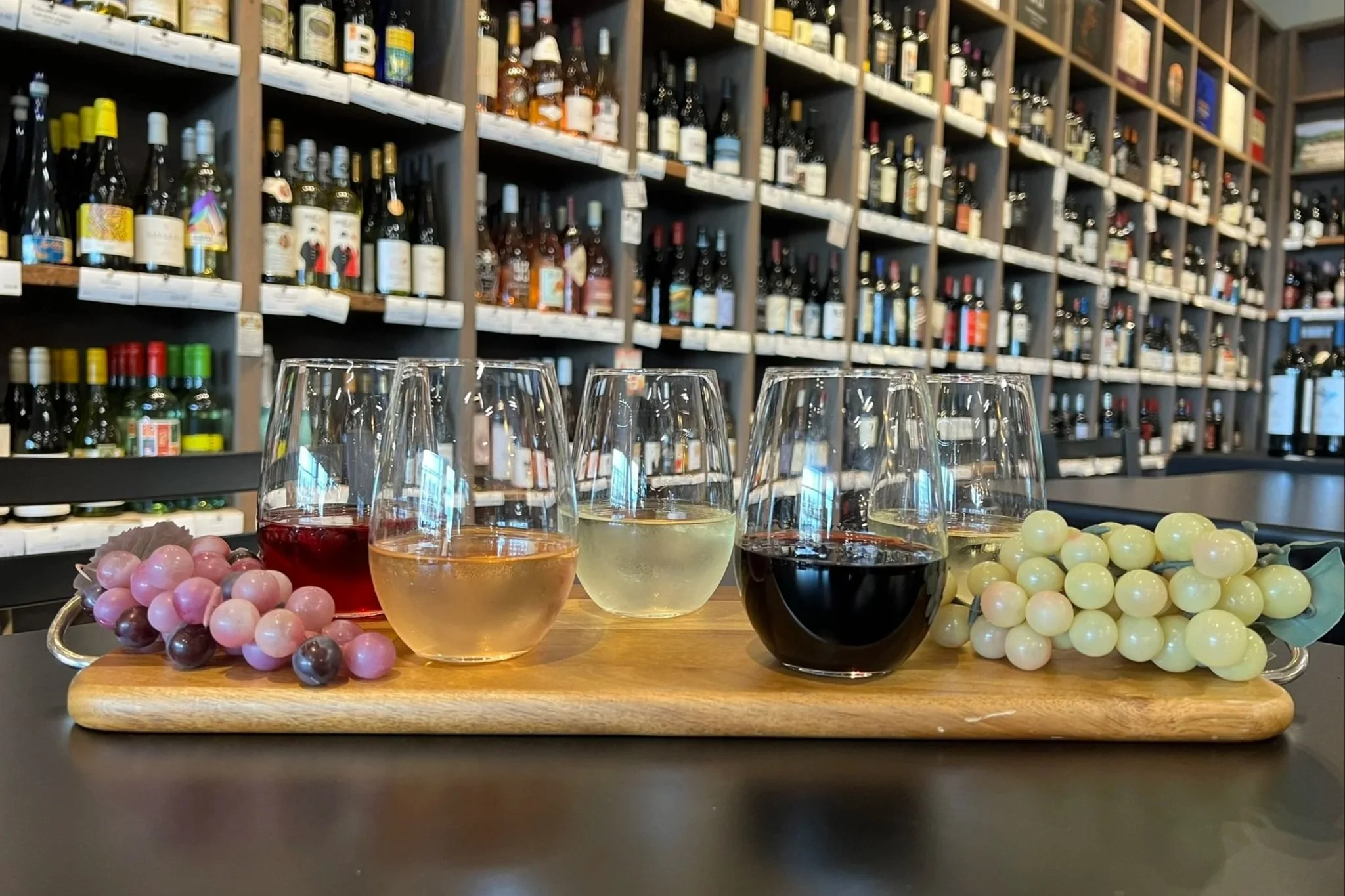 A wooden tray with four glasses of different types of wine, surrounded by bunches of grapes, on a table in front of a wine shelf filled with bottles.