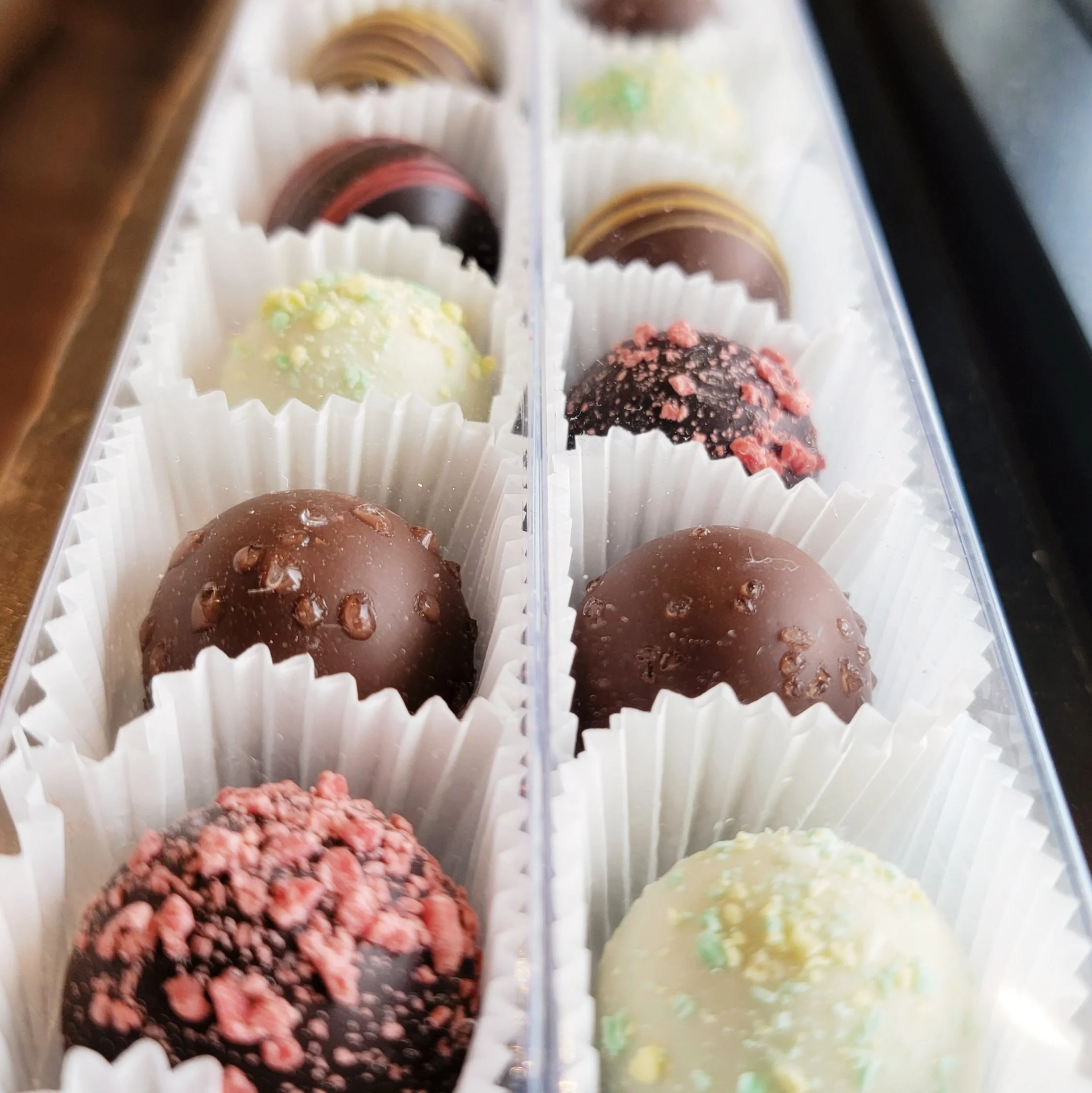 Close-up of a plastic tray filled with assorted truffles or chocolate confections, each in a paper cup, with some topped with sprinkles or crushed toppings.