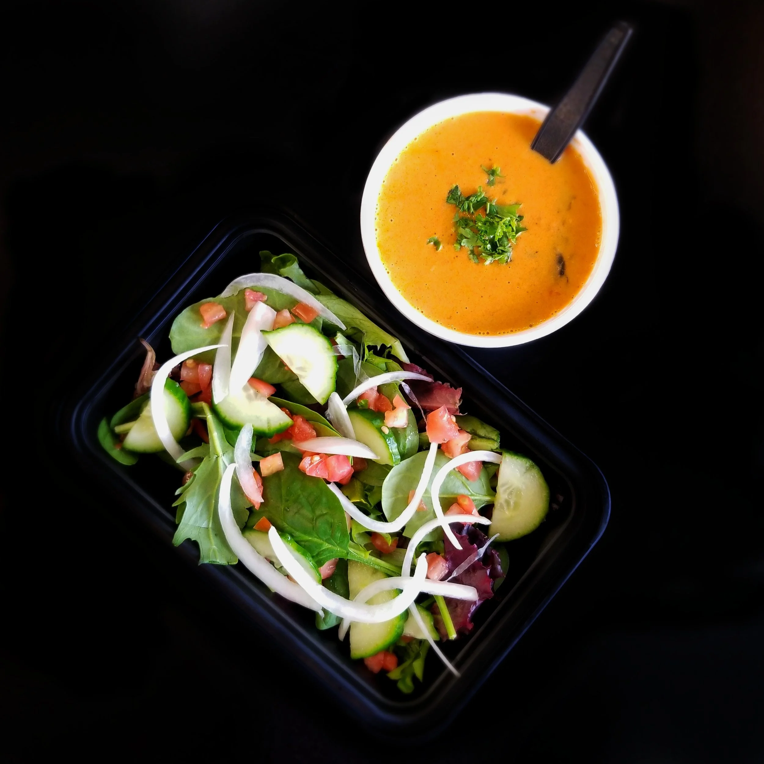 A bowl of orange soup garnished with chopped green herbs, served with a side salad of mixed greens, sliced cucumbers, cherry tomatoes, onions, and lemon slices on a black background.