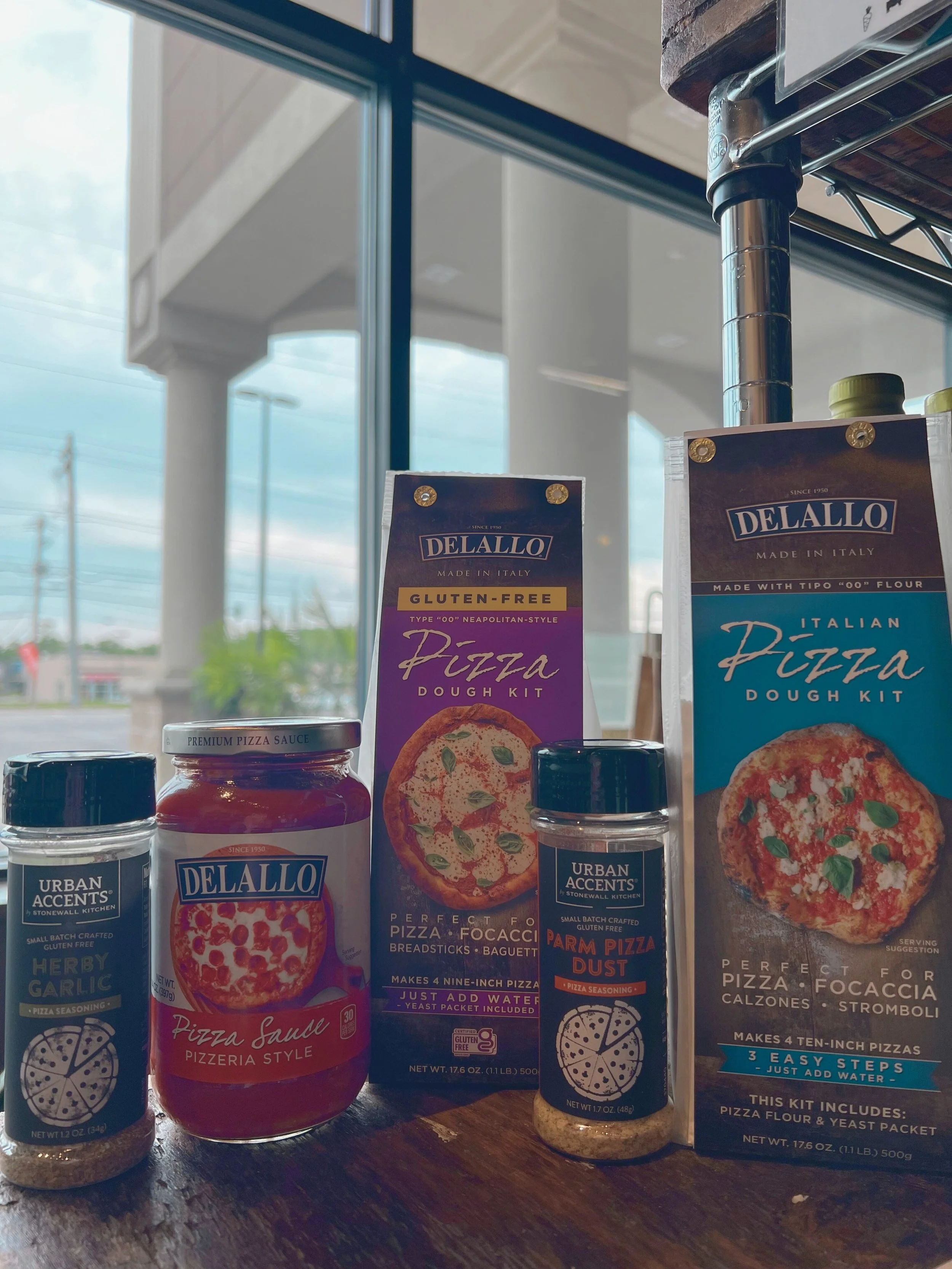 Various pizza dough kits and sauces on a wooden table inside a store with large windows.