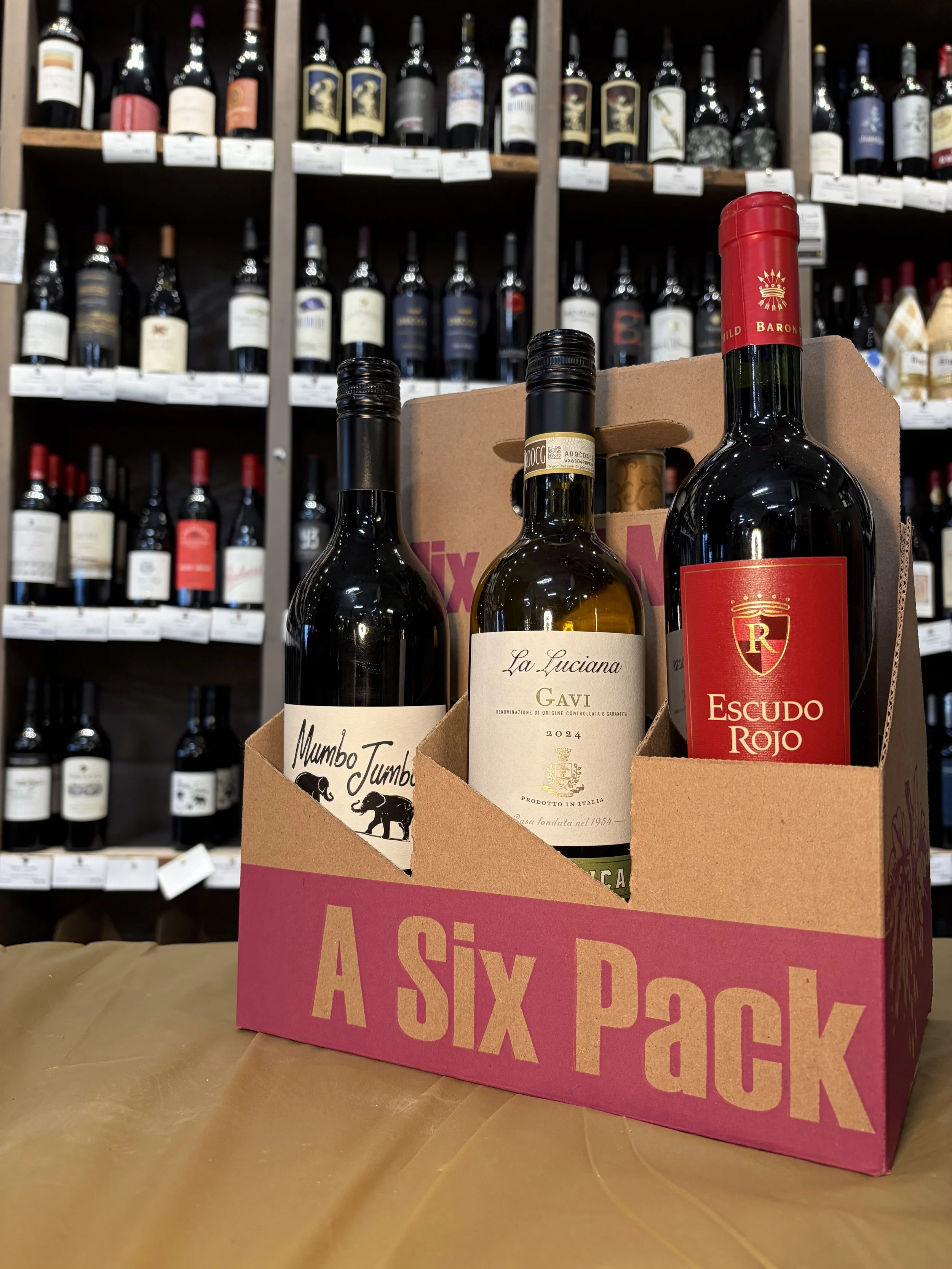 A six-pack of wine bottles in a pink cardboard carrier. The bottles are placed on a wooden surface with a background of shelves filled with many more bottles of wine.
