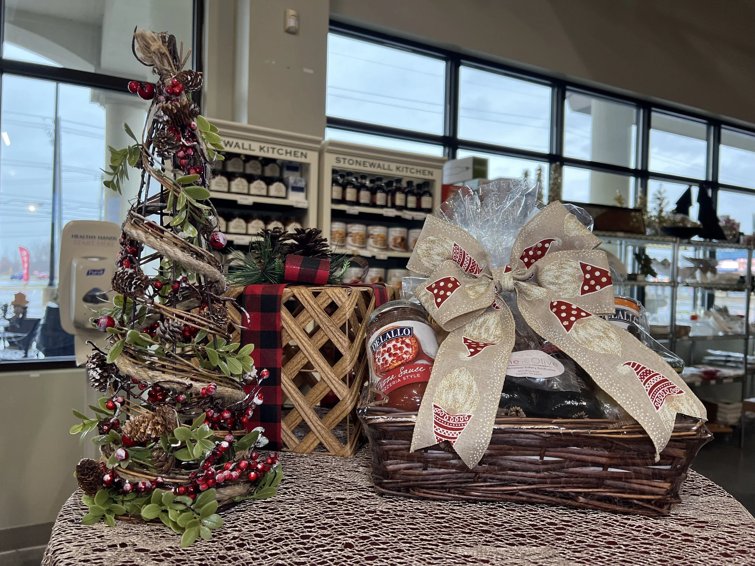 Holiday-themed decorations including a small decorative Christmas tree with pinecones, berries, and green leaves, placed in a basket with a red and black plaid ribbon. There is lace and a large bow with red and white polka dots next to the basket. In the background, store shelves with jars and products are visible.
