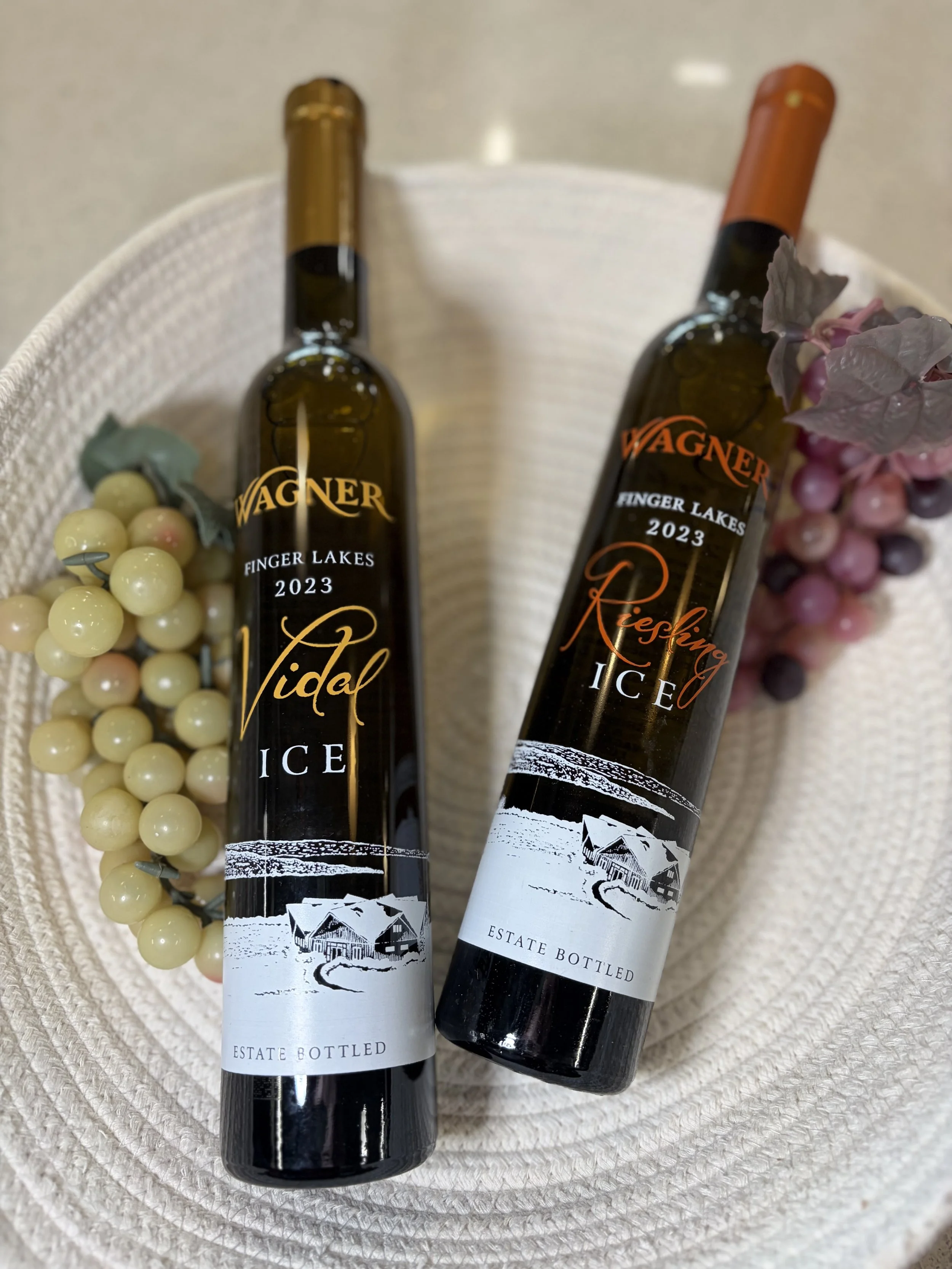 Two bottles of wine with labels reading 'Finger Lakes 2023 Riesling' and an illustration of a winery, placed on a woven white surface with purple and white grapes and white berries and greenery nearby.