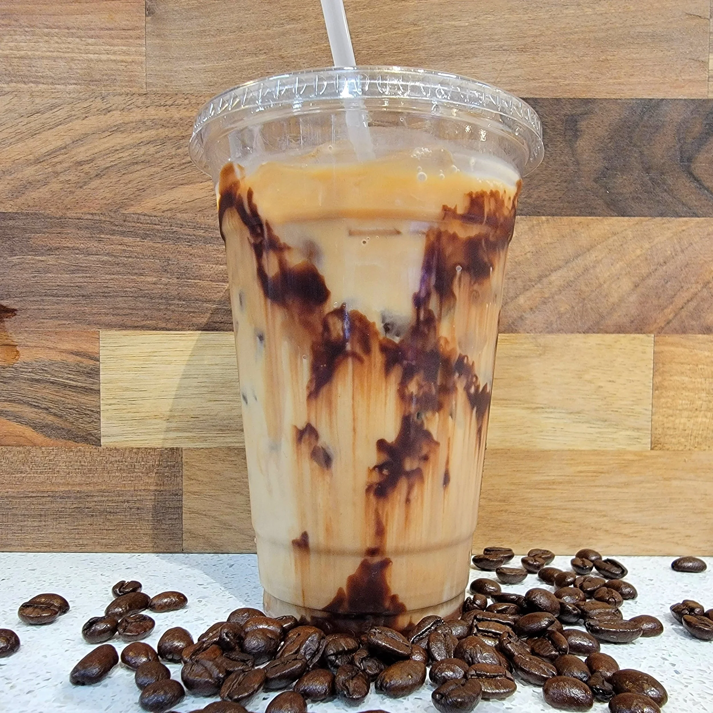 A plastic cup filled with iced coffee, with cream and chocolate syrup swirls, on a white speckled countertop, surrounded by scattered coffee beans, against a wooden background.