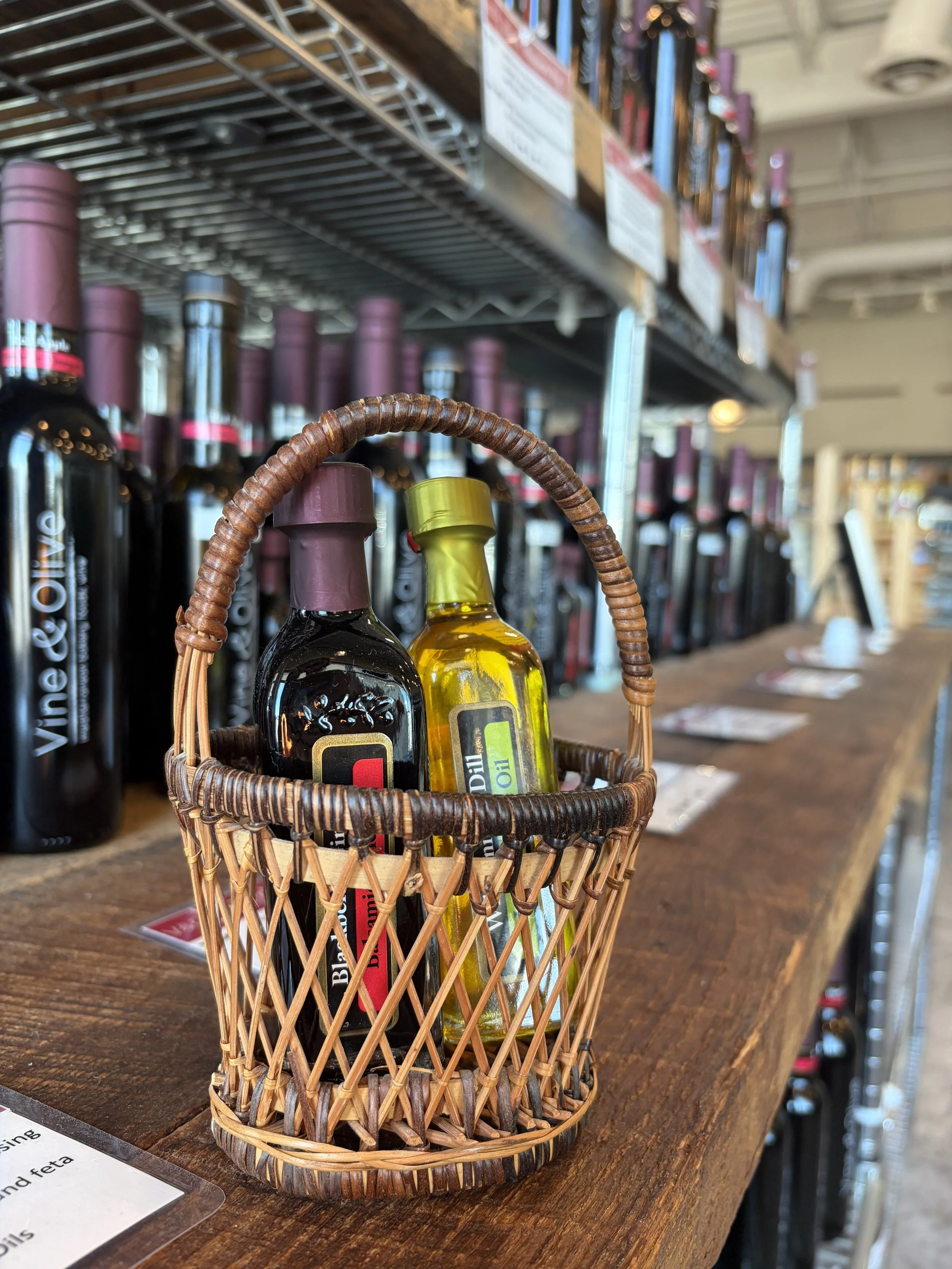 Small wicker basket with two bottles filled with oil or vinegar, set on a wooden table in a store with shelves of other bottles in the background.