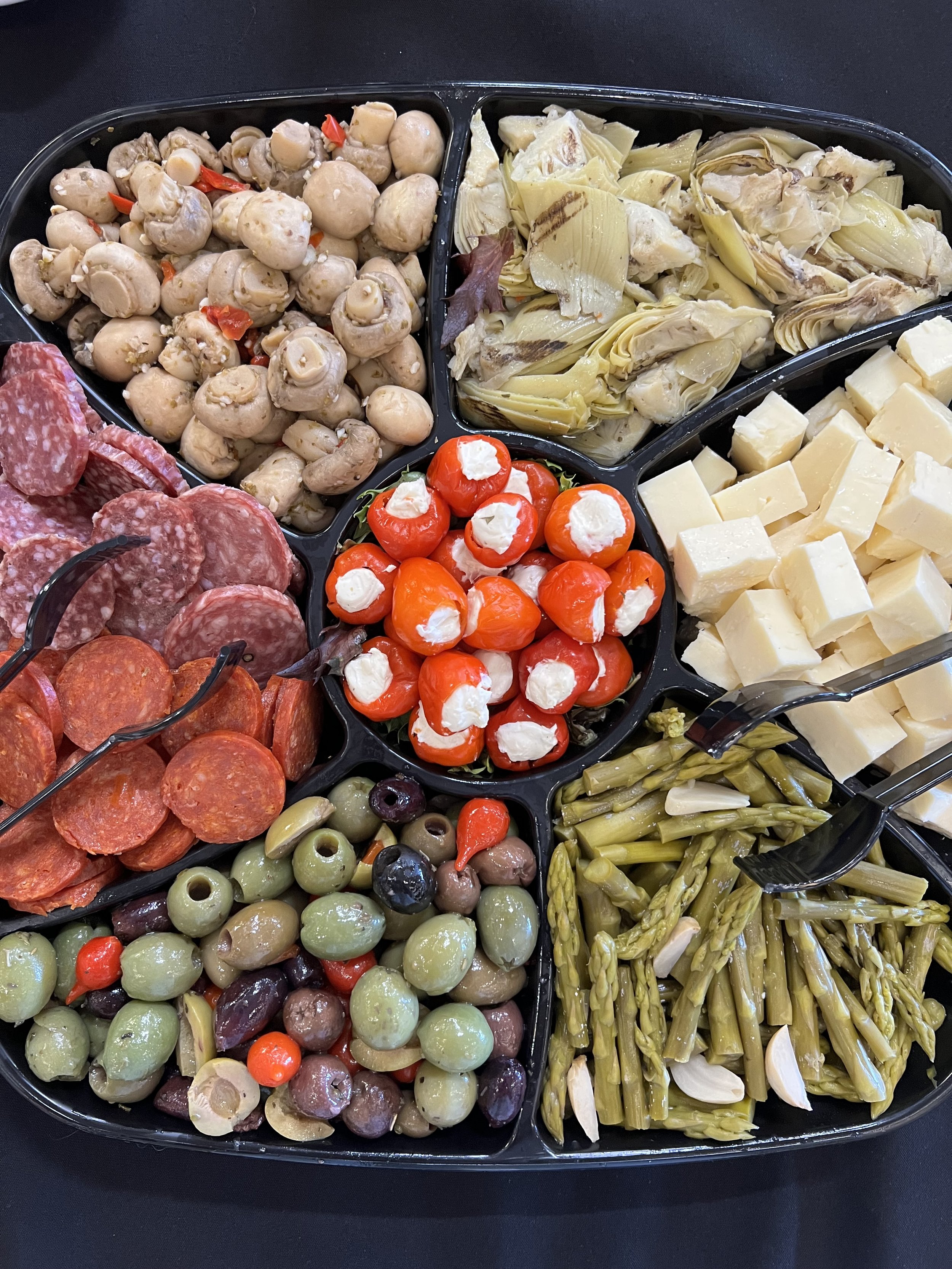 Assorted appetizer tray with marinated mushrooms, artichoke hearts, cherry tomatoes with mozzarella, mixed olives, sliced salami, cubed cheese, and cooked green vegetables on a black surface.