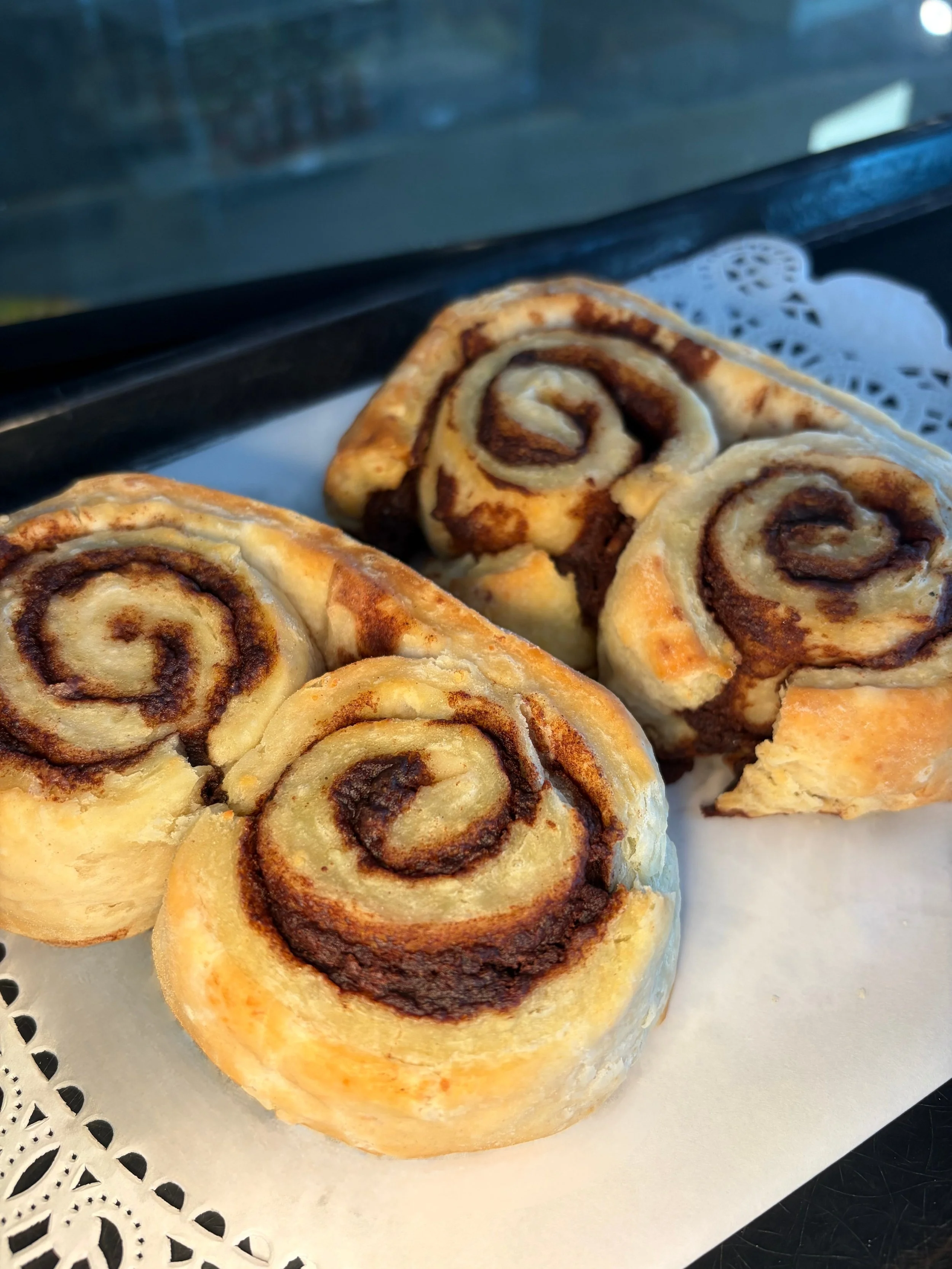 Close-up of cinnamon roll pastries with swirls of cinnamon sugar filling, placed on a decorative paper doily on a tray.