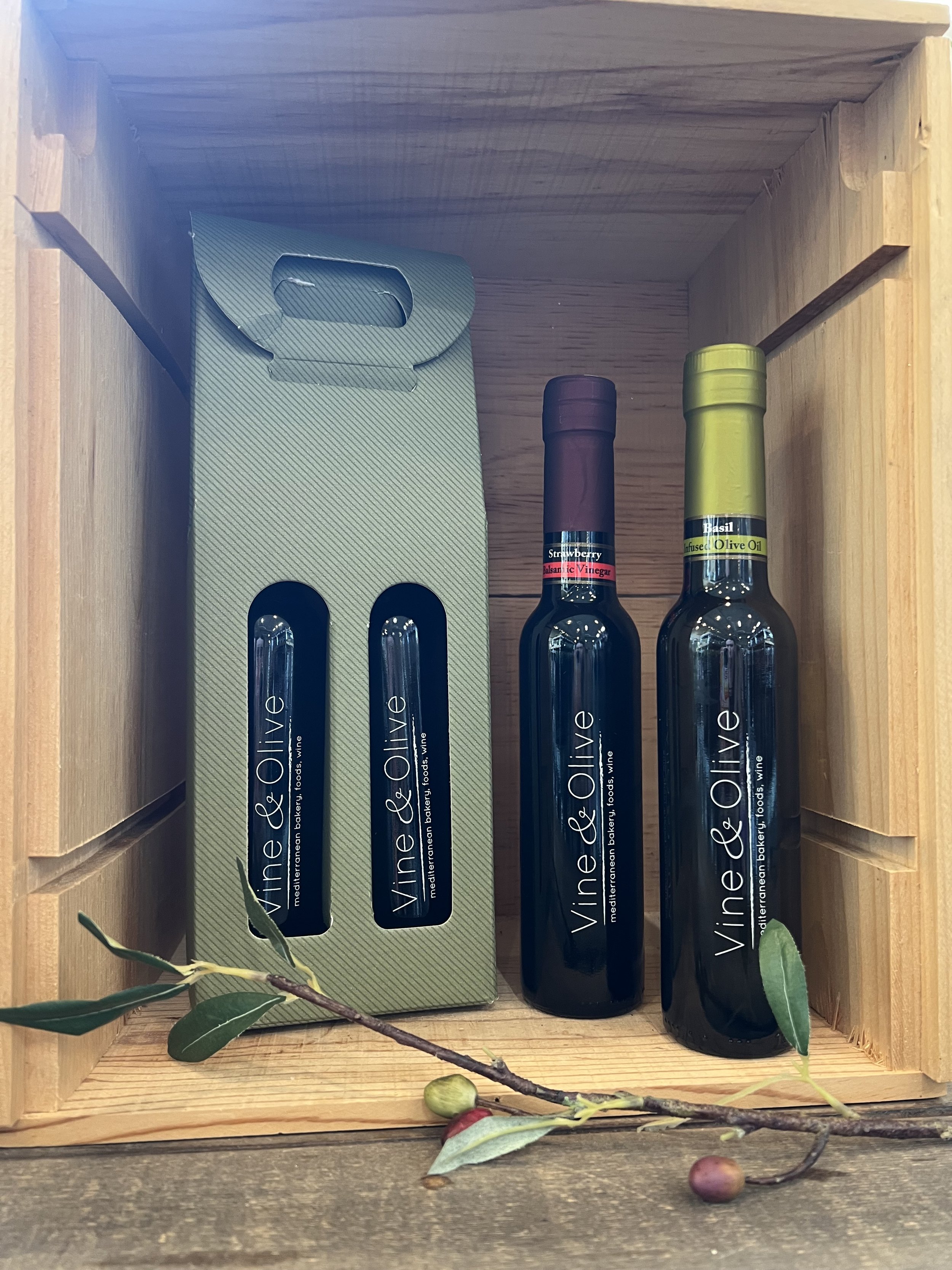 A wooden box with bottles of olive oil labeled 'Vine & Olive' inside, with some olive branches and olives placed in front of the box.