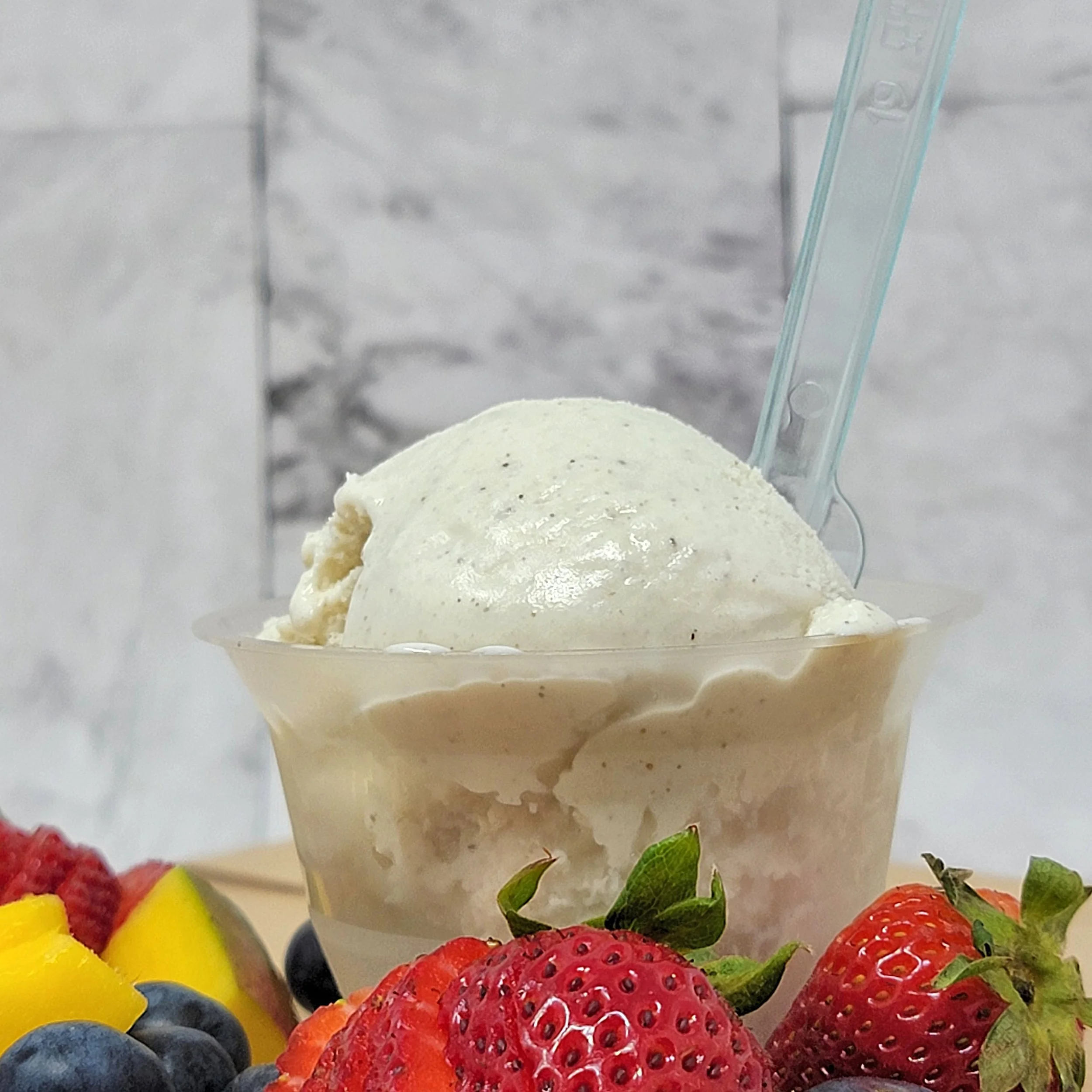 Close-up of a cup with gelato topped with strawberries, blueberries, and green leaves, with a blurred background of other fruits.