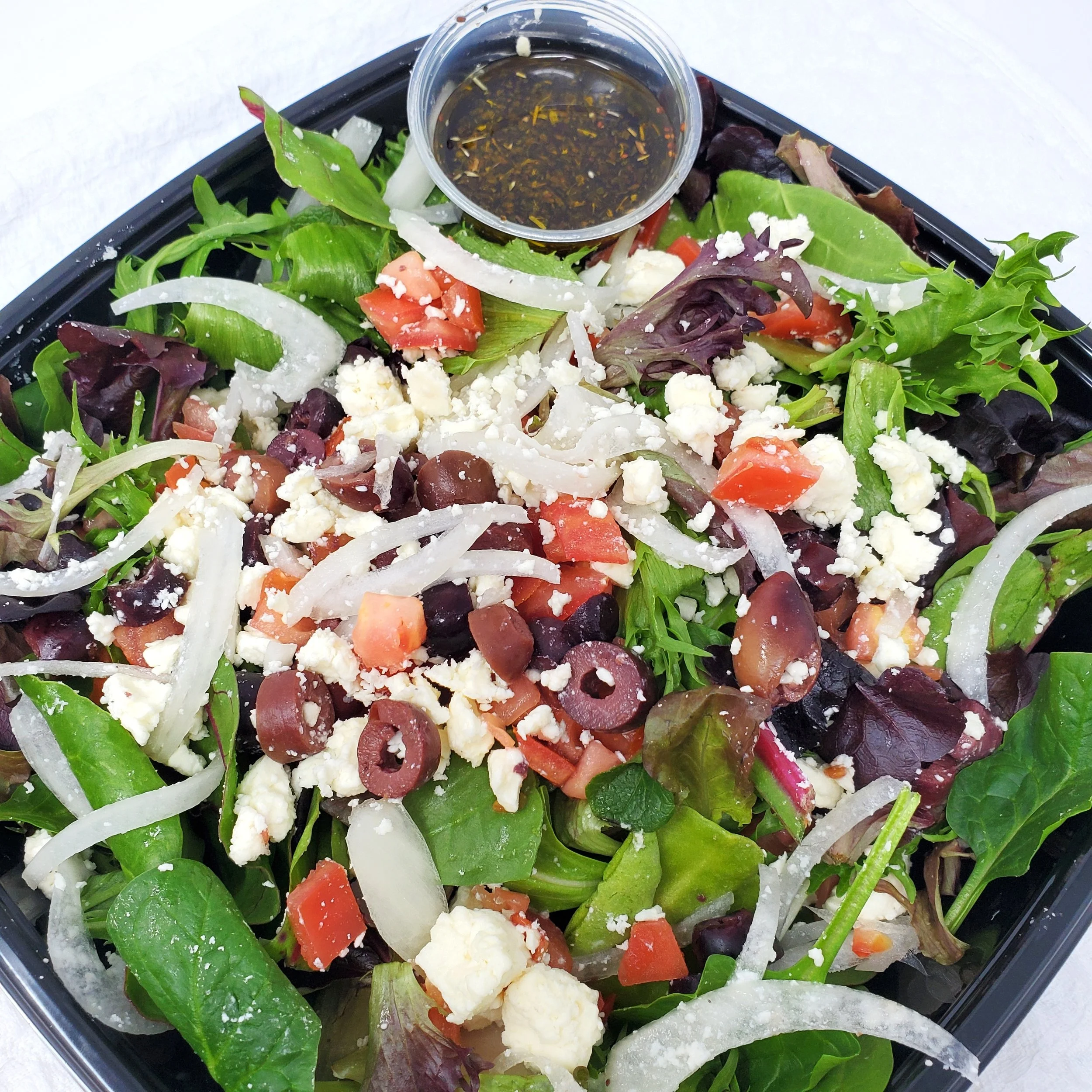 Mixed green salad with cherry tomatoes, sliced onions, crumbled feta cheese, black olives, and a side of balsamic vinaigrette dressing in a small cup.