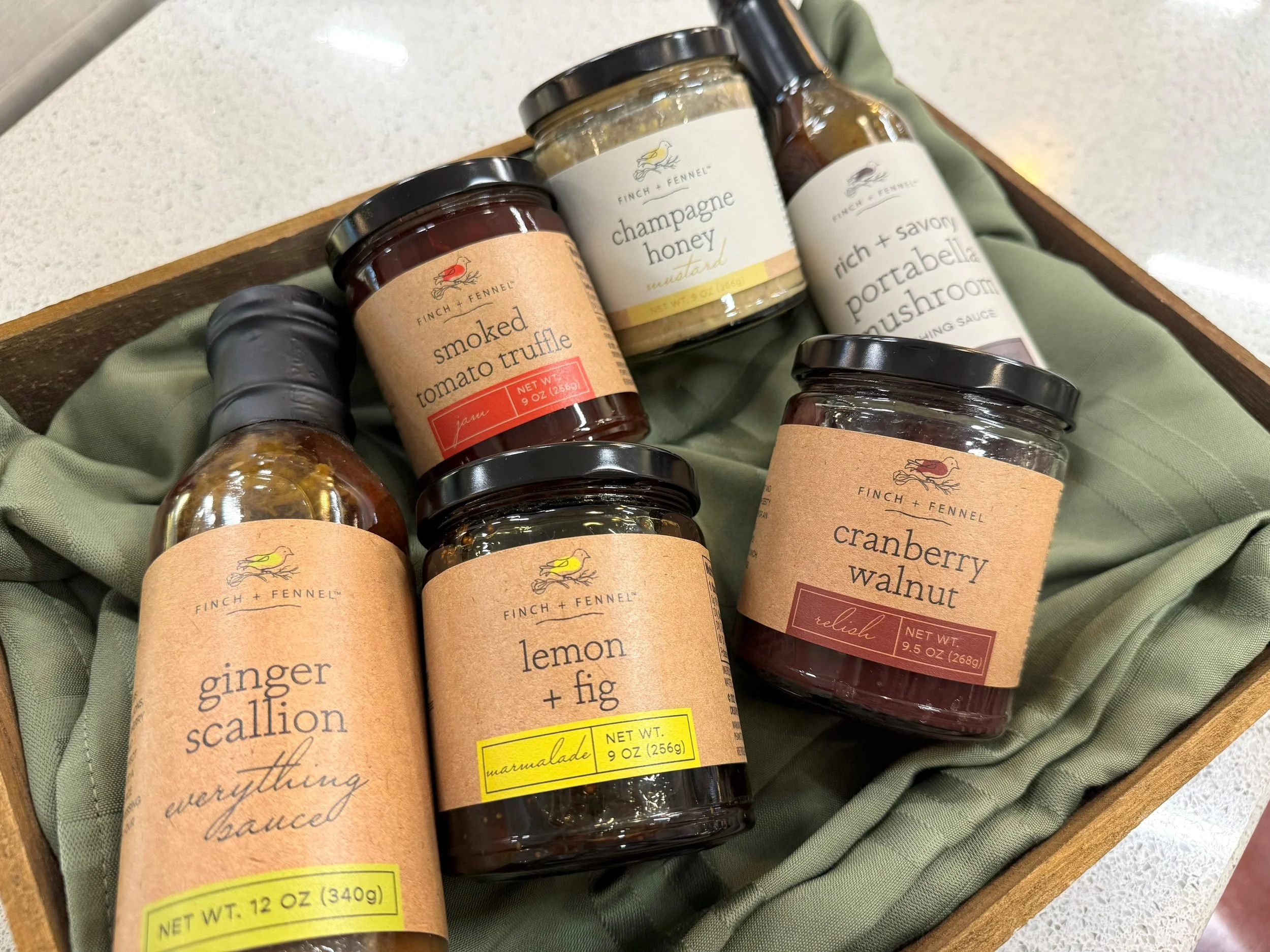 A wooden tray lined with green fabric containing six jars of Finch + Fennel spreads and sauces, including tomato truffle jam, champagne honey mustard, and cranberry walnut relish.