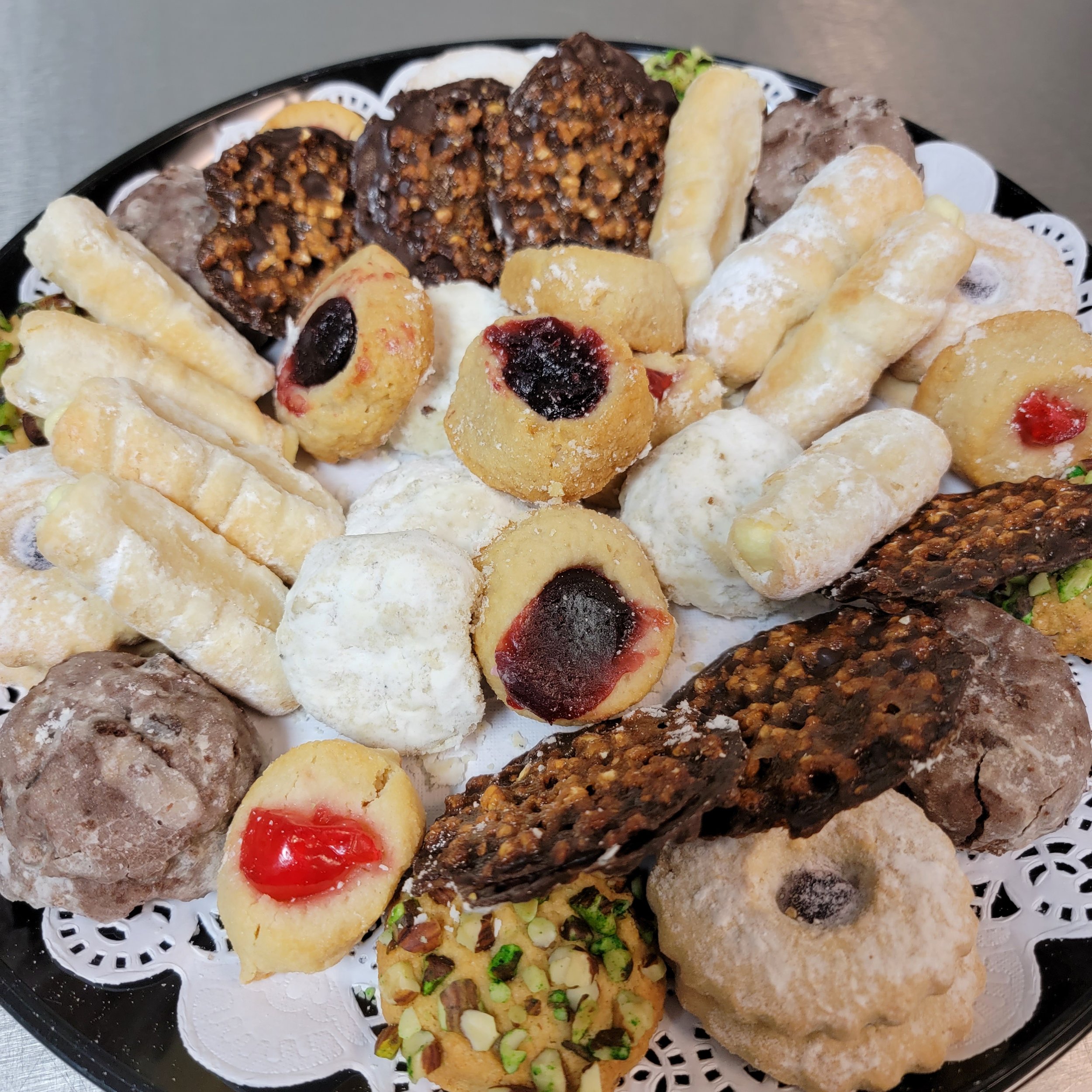 An oval black tray filled with assorted bite-sized cookies and confections, including chocolate-covered nut clusters, fruit-filled cookies, and powdered sugar-coated treats arranged on a decorative paper doily.