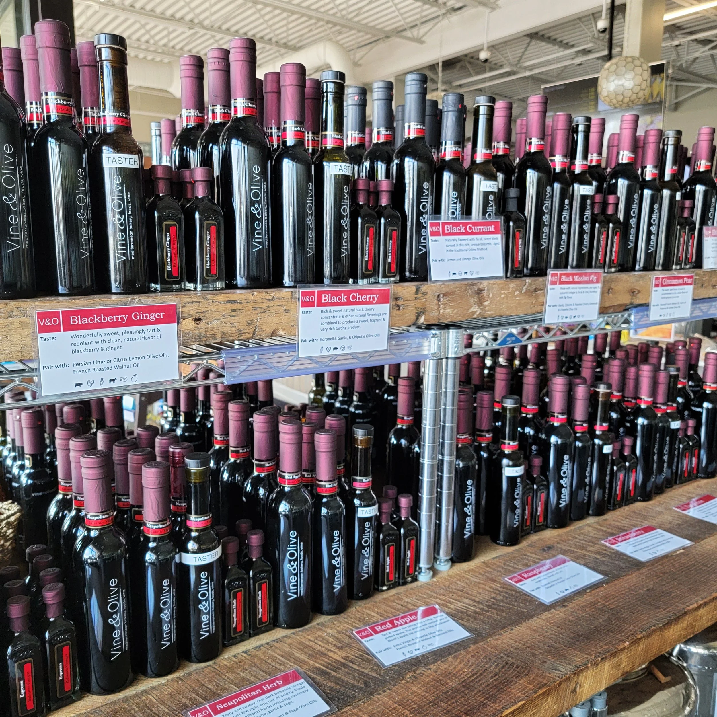 Rows of dark glass bottles of vinegar with pink and black caps on a wooden display shelf in a grocery store, labeled with various flavors like blackberry ginger and black cherry.