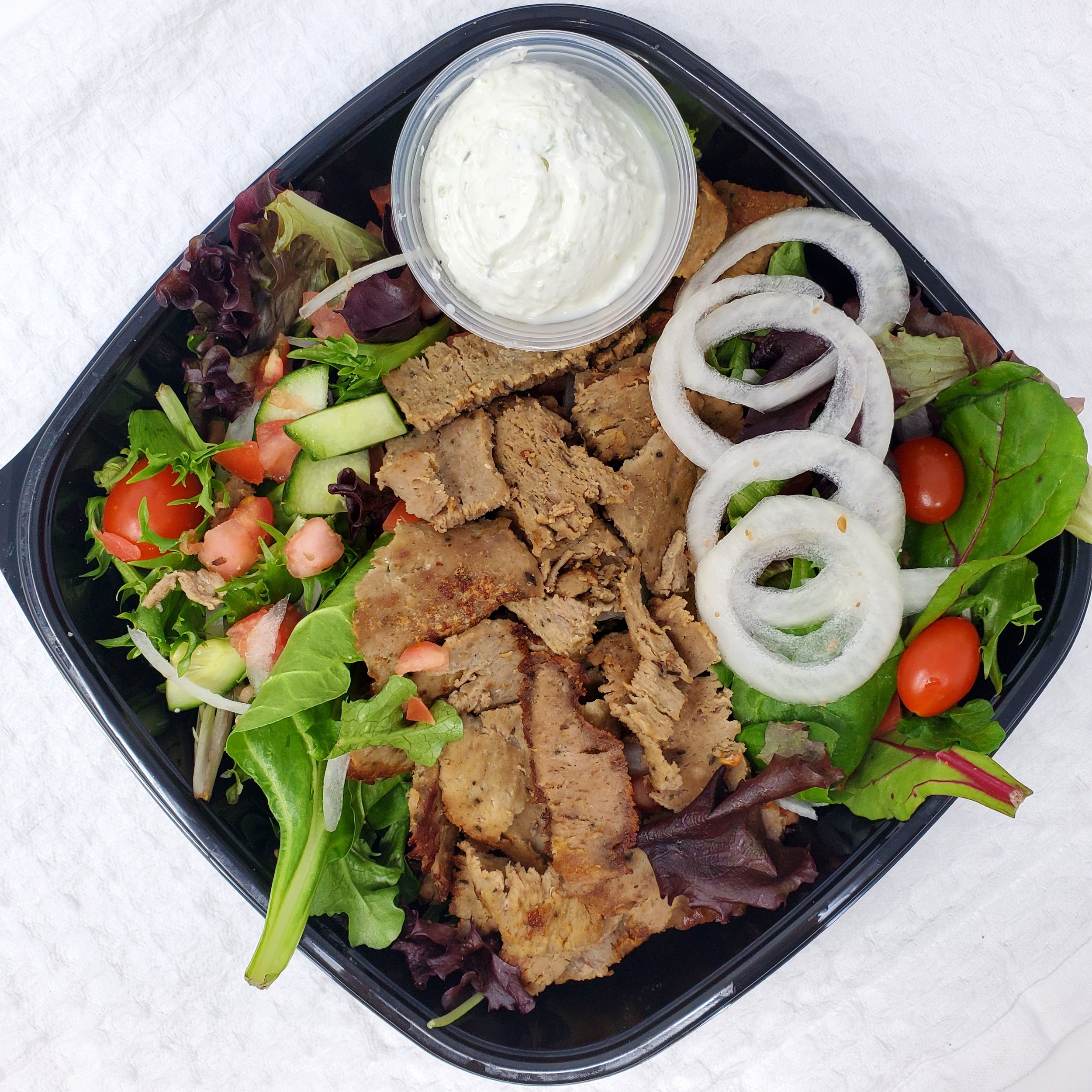 A black plastic takeout container filled with a salad containing leafy greens, cherry tomatoes, cucumber slices, and slices of grilled chicken, topped with a small container of ranch dressing and sliced onion rings.