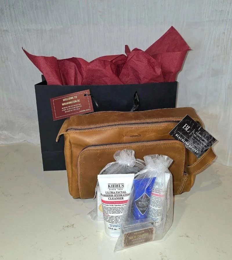 A black bag with a red tag, a brown leather toiletry bag, and a clear plastic bag with skincare products, all placed against a light-colored textured background.