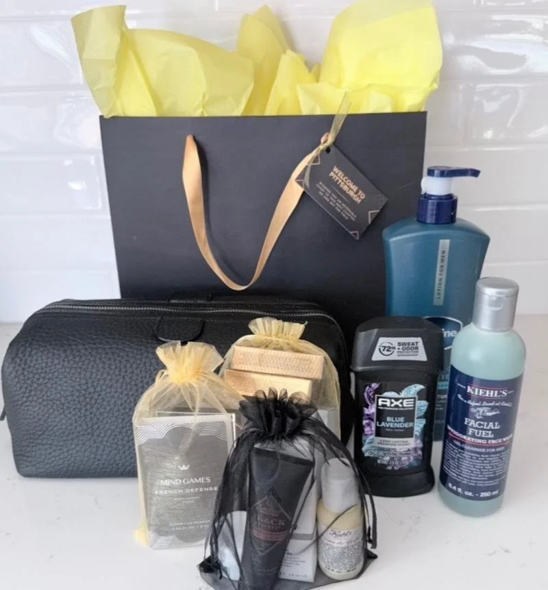 A collection of personal care and grooming products including body wash, facial cleanser, deodorant, and lotions, along with a black toiletry bag, and a black gift bag with yellow tissue paper, all arranged on a white surface against a white tiled wall.