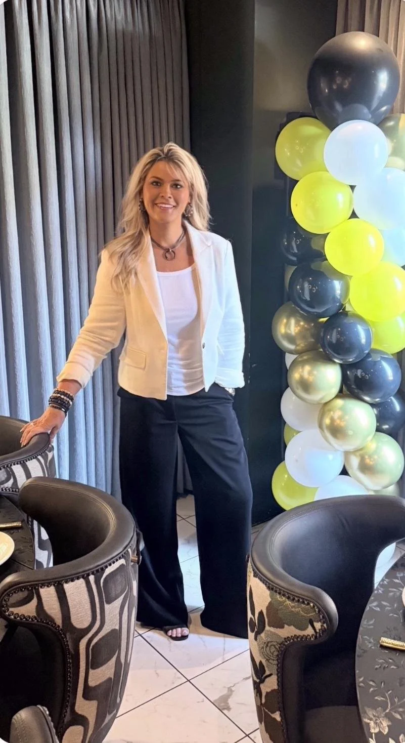 A woman dressed in a white blazer, white top, and dark pants standing beside a table with chairs, celebrating indoors with a balloon decoration in black, white, yellow, and metallic gold.