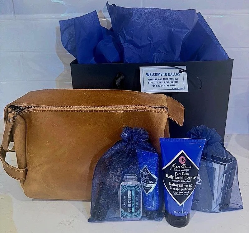 A brown leather makeup bag with a zipper, two small blue gift bags with dark blue tissue paper inside, a black envelope with a welcome message, and a tube of Jack Black pure clean daily facial cleanser.
