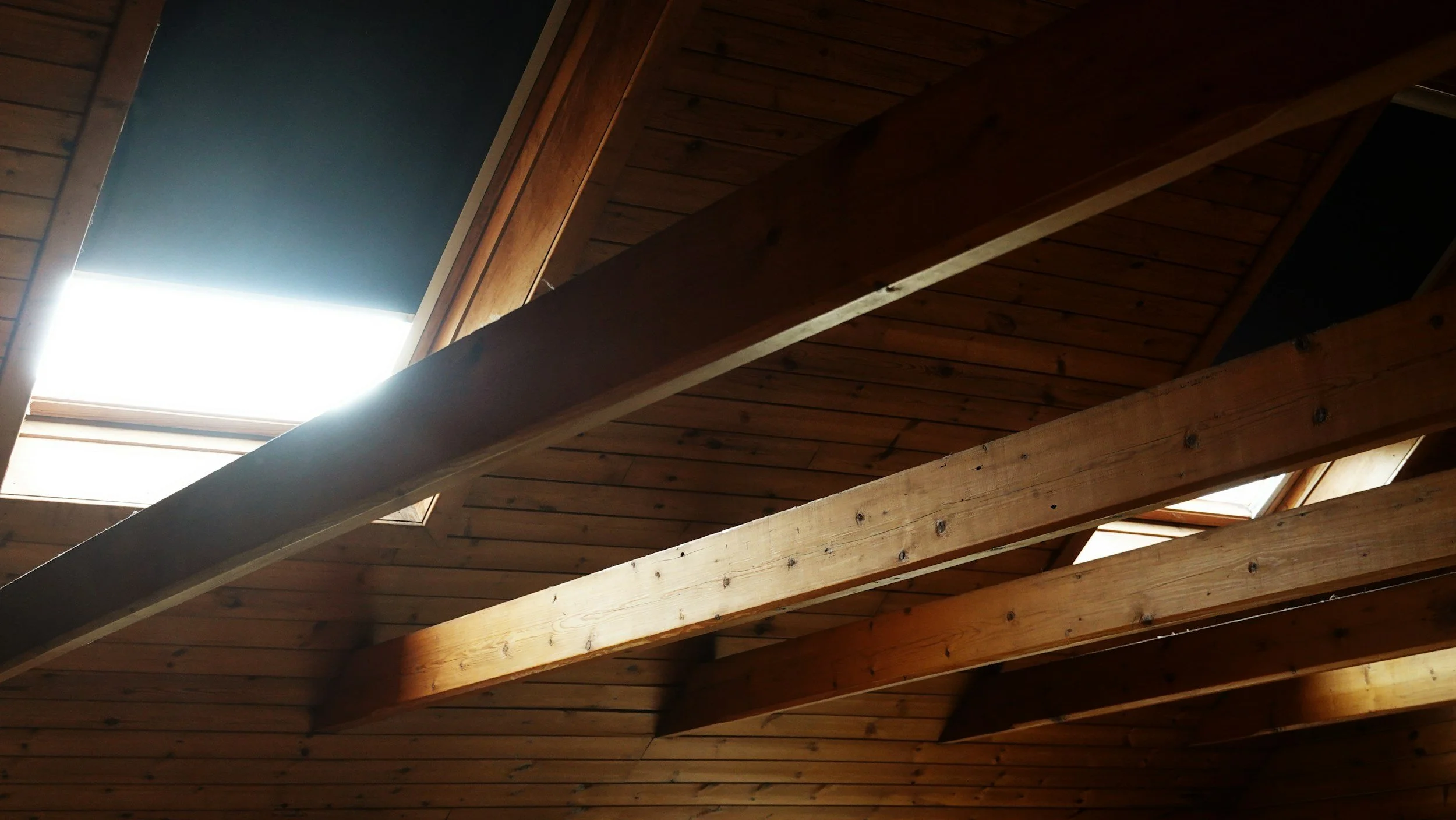 Photo of a sloped wooden ceiling with exposed beams, featuring skylights that allow natural light to shine through.