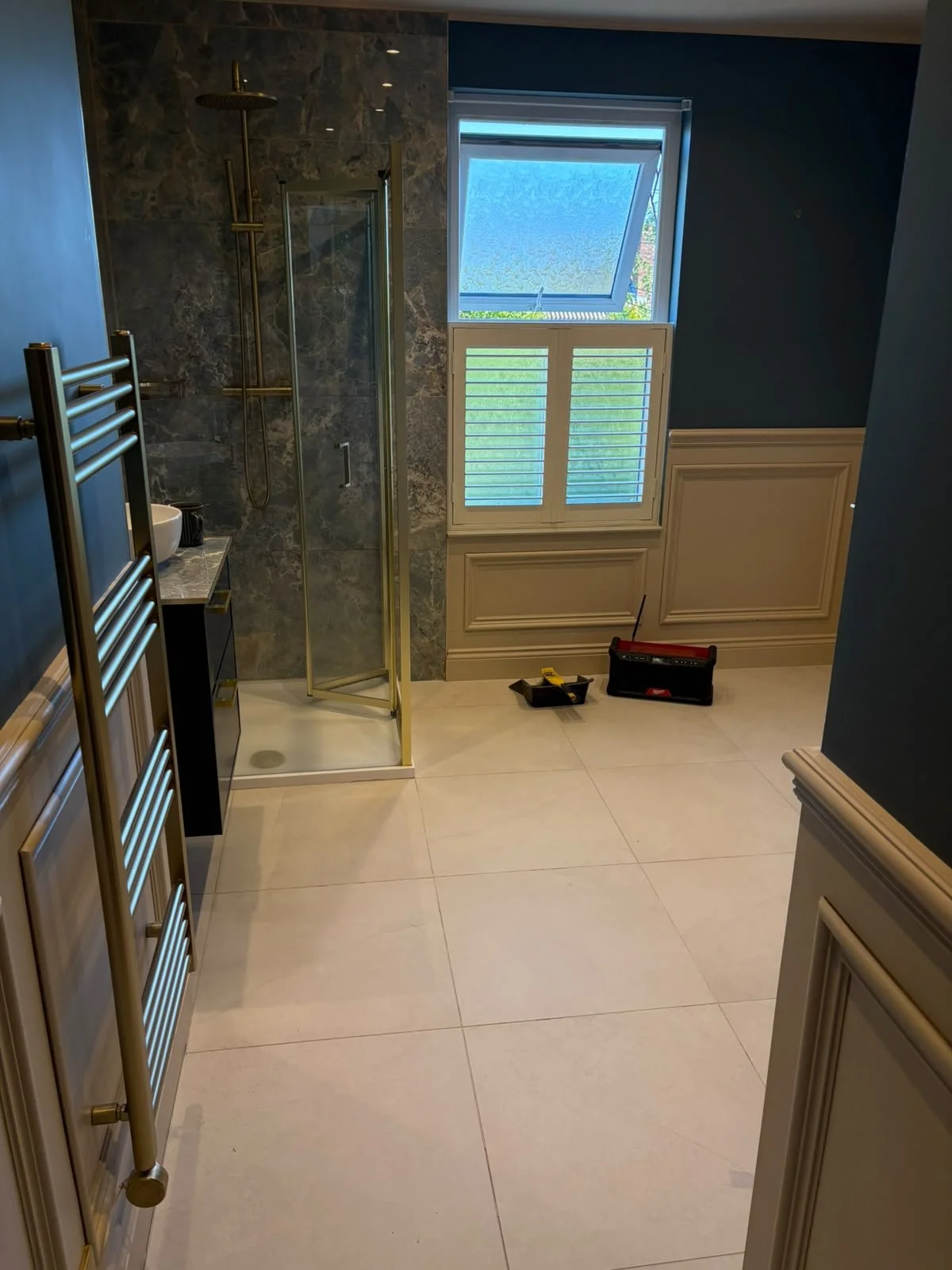 A bathroom with a glass shower enclosure, a window with shutters, a towel radiator, and tools on the floor indicating ongoing installation or repairs.