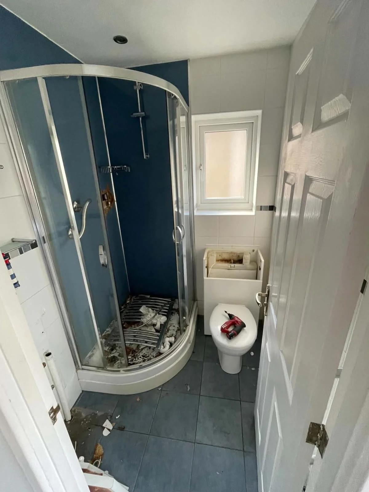 A small bathroom in the process of cleaning. Features a corner shower with a blue interior, a window on the wall, a toilet with a cordless drill on top, and debris and dirty towels on the floor and inside the shower. The door is open, showing the roo