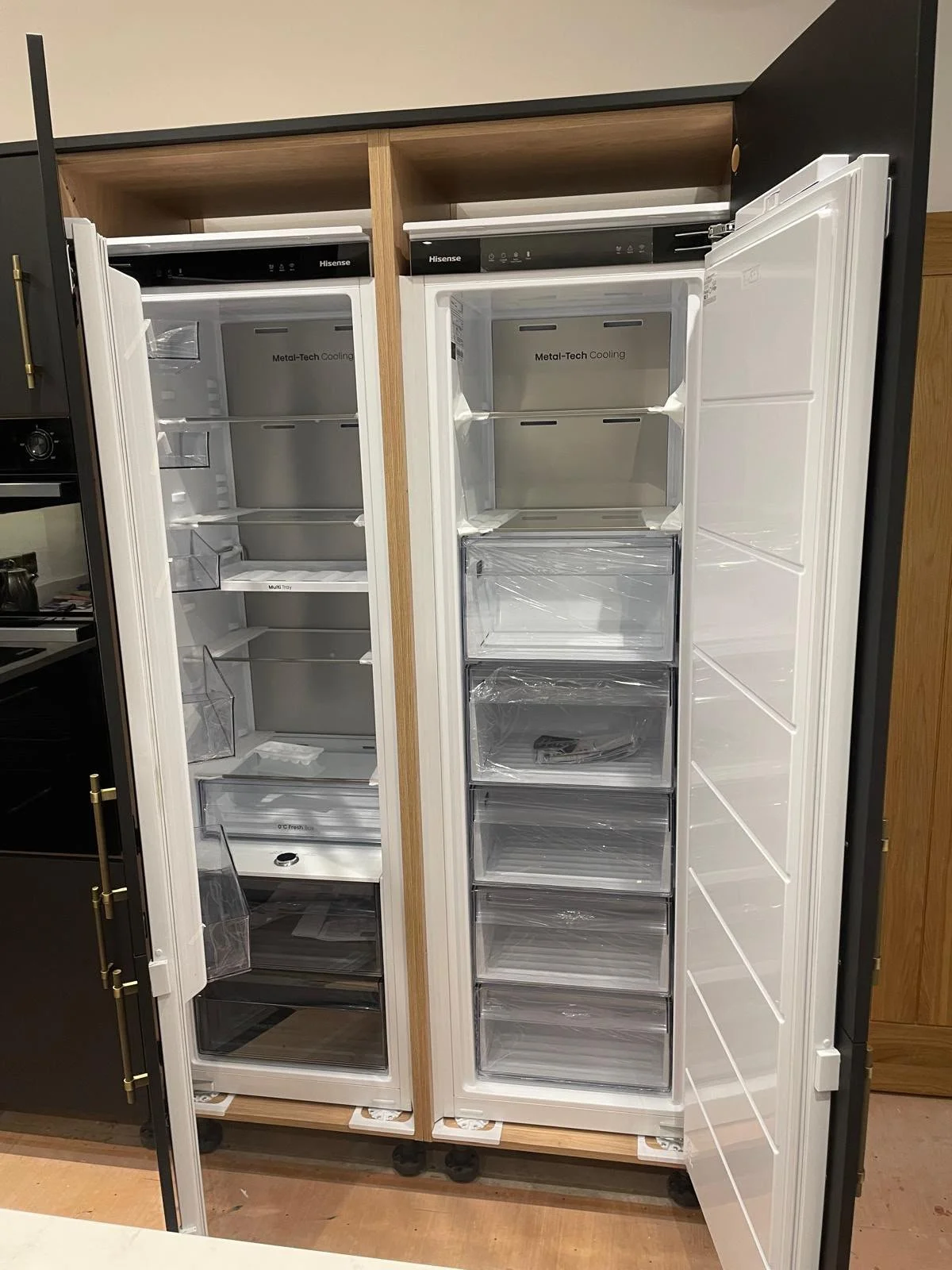 Open door refrigerator with empty shelves and drawers, side-by-side with another empty refrigerator, both placed on a wooden stand.