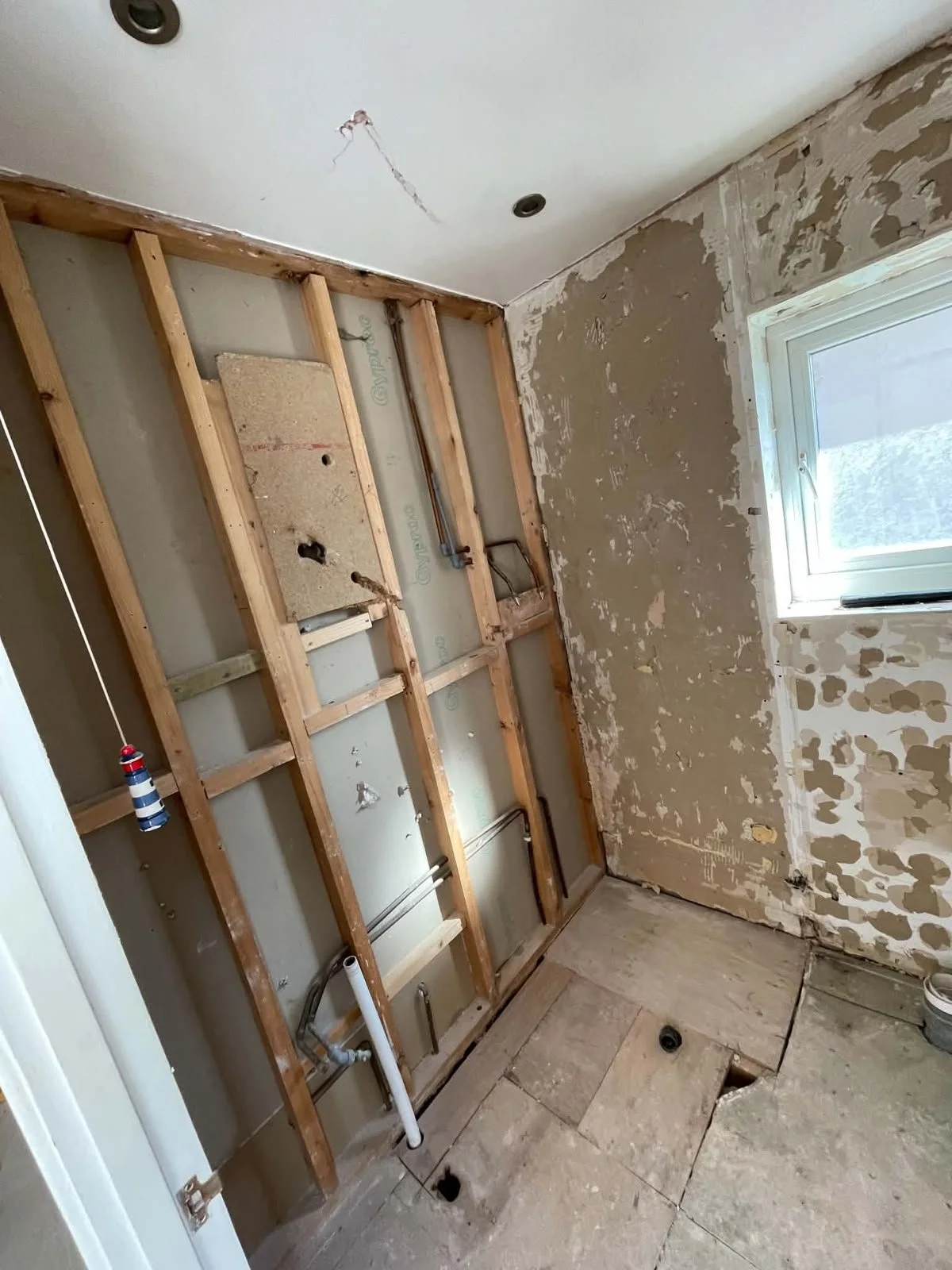 Room under renovation with exposed wooden framing, plumbing pipes, and drywall partially removed, revealing wall studs and plumbing, with a window on the right wall.