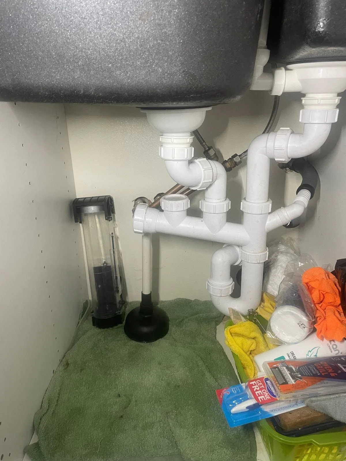 Underneath sink plumbing with white PVC pipes, drain trap, and water supply lines. Various tools and cleaning supplies are on the green towel and nearby surface.